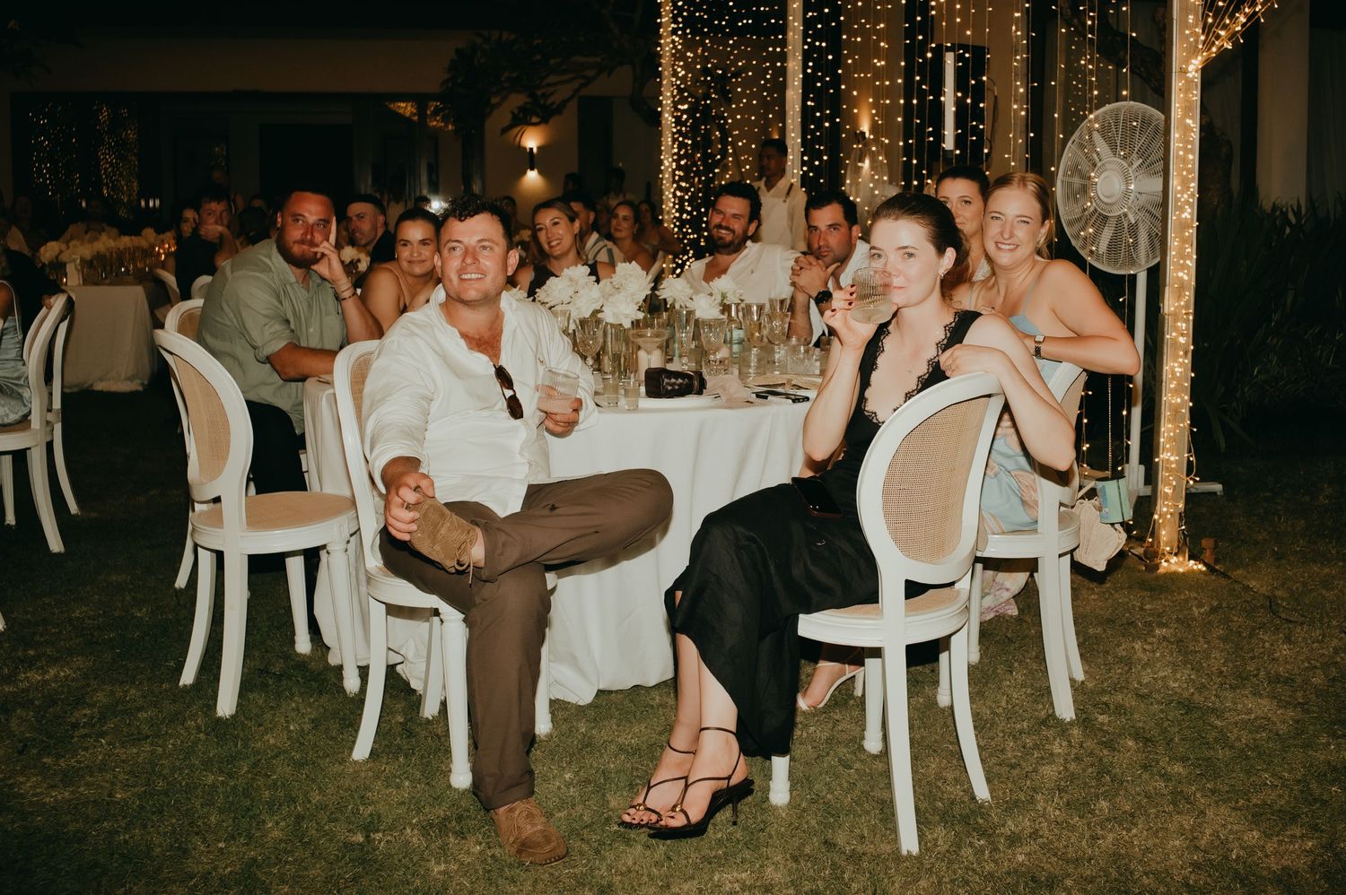 Wedding guests seated outdoors at night, smiling and enjoying the reception under fairy lights.