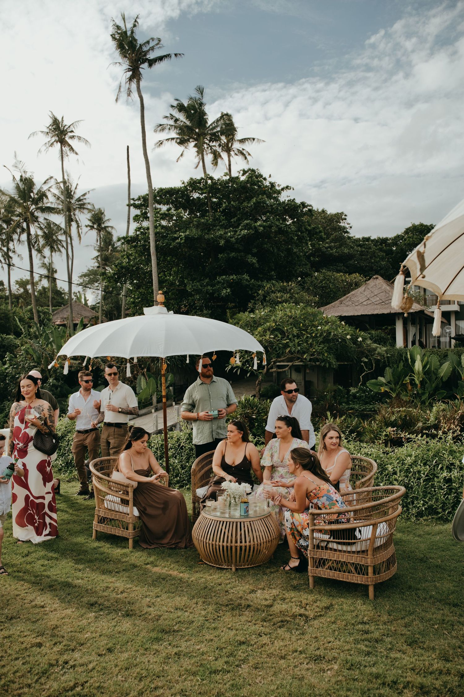 Guests mingle at a tropical outdoor wedding cocktail hour with rattan furniture and a white Balinese umbrella.