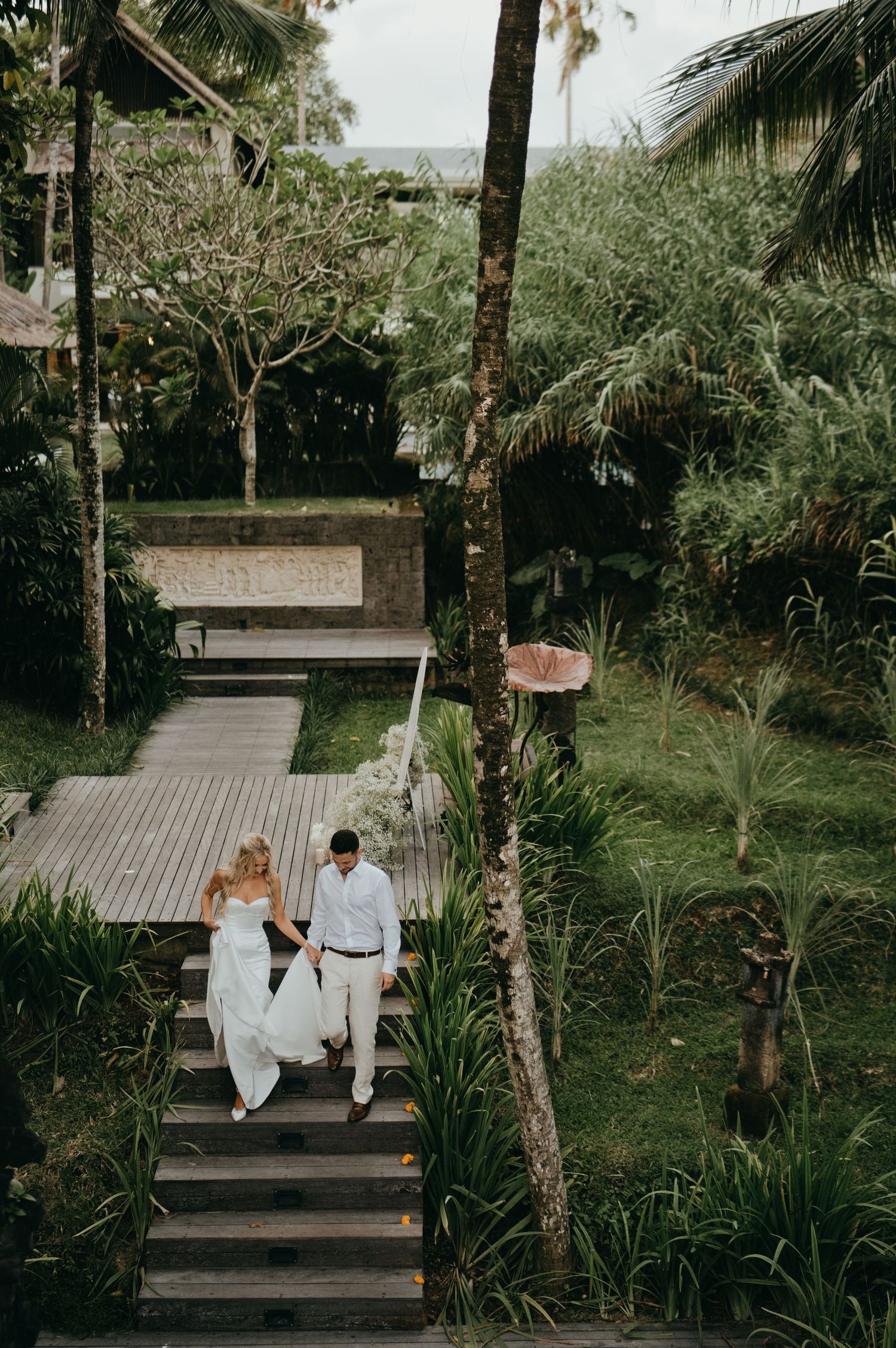Aerial view of bride and groom walking down wooden steps through lush tropical garden.