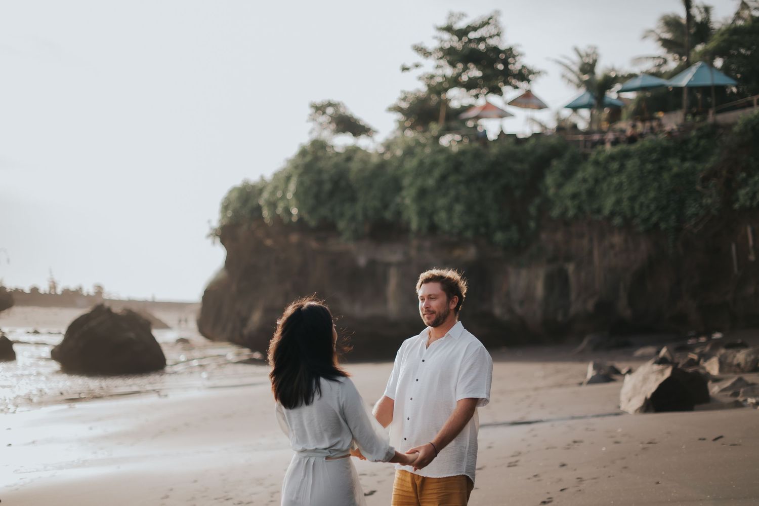 Couple walking hand in hand on a scenic beach with rocky cliffs and palm trees at sunset.