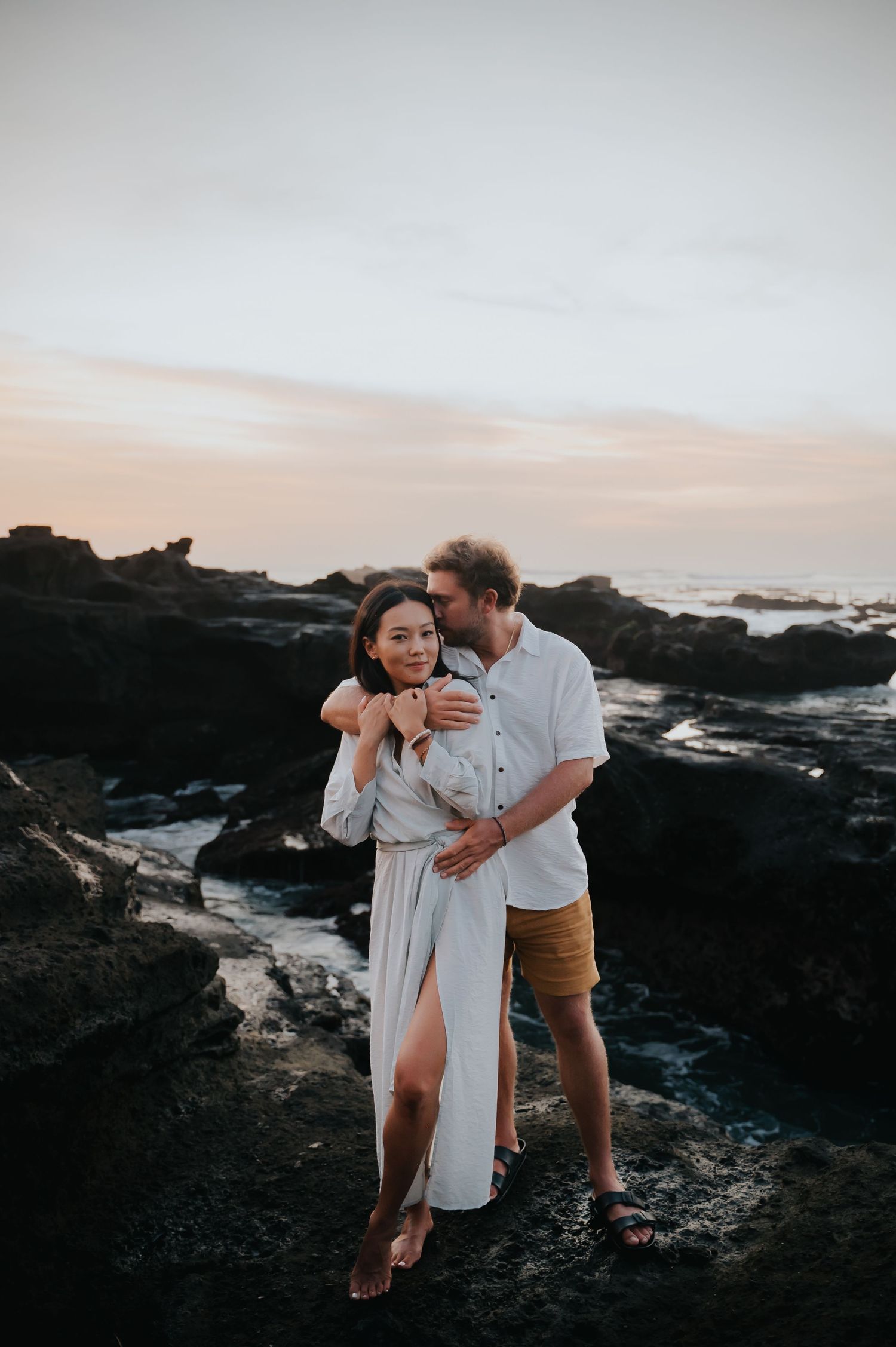 A couple shares an intimate moment on rocky coastal cliffs at sunset with ocean waves crashing in the background.