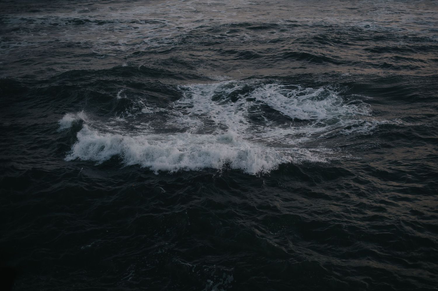 Dark ocean waves roll and crash against each other under an overcast sky creating a moody seascape.