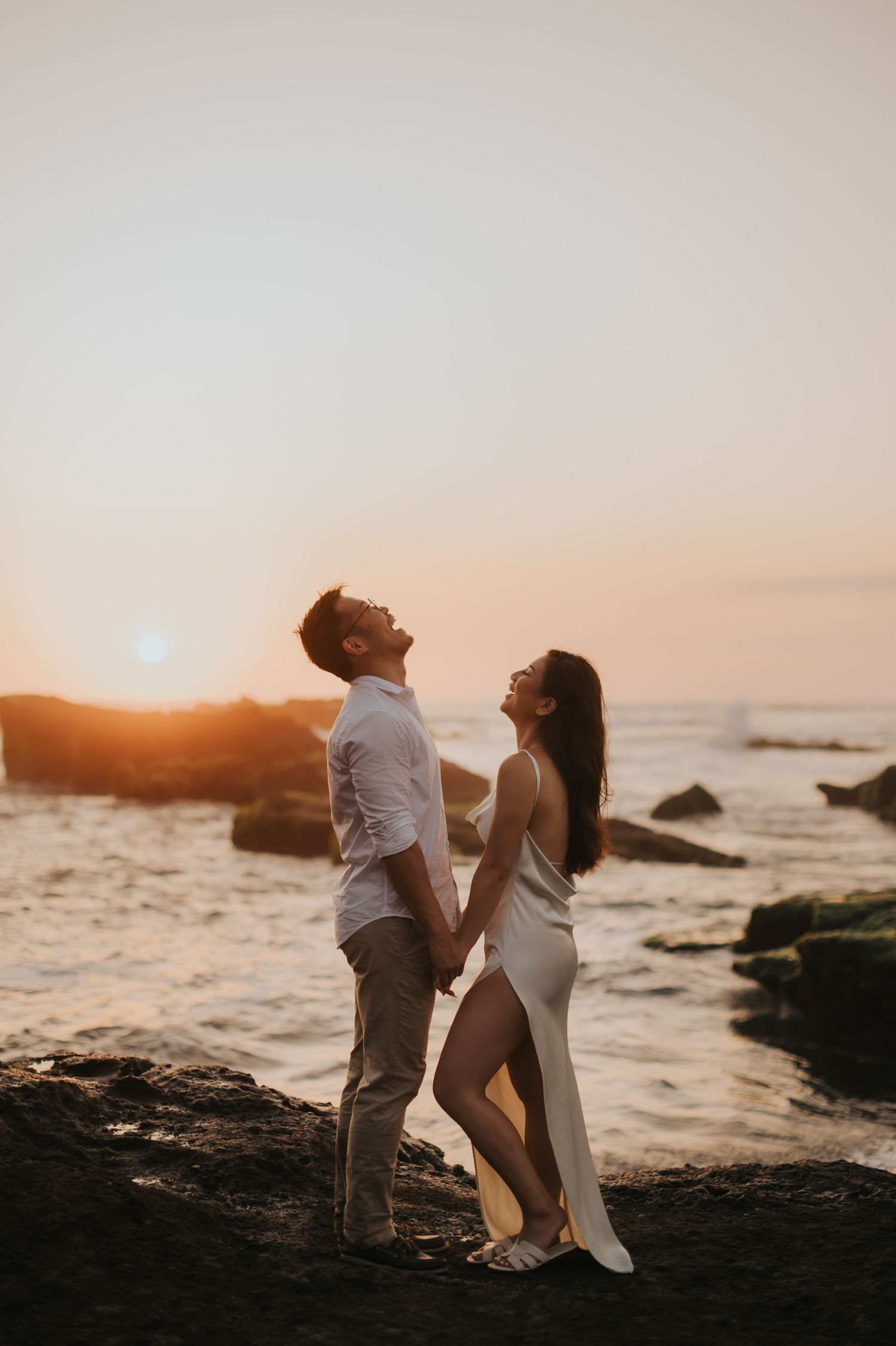 Couple holding hands on rocky beach at golden sunset, woman in white dress and man in white shirt gazing upward romantically.