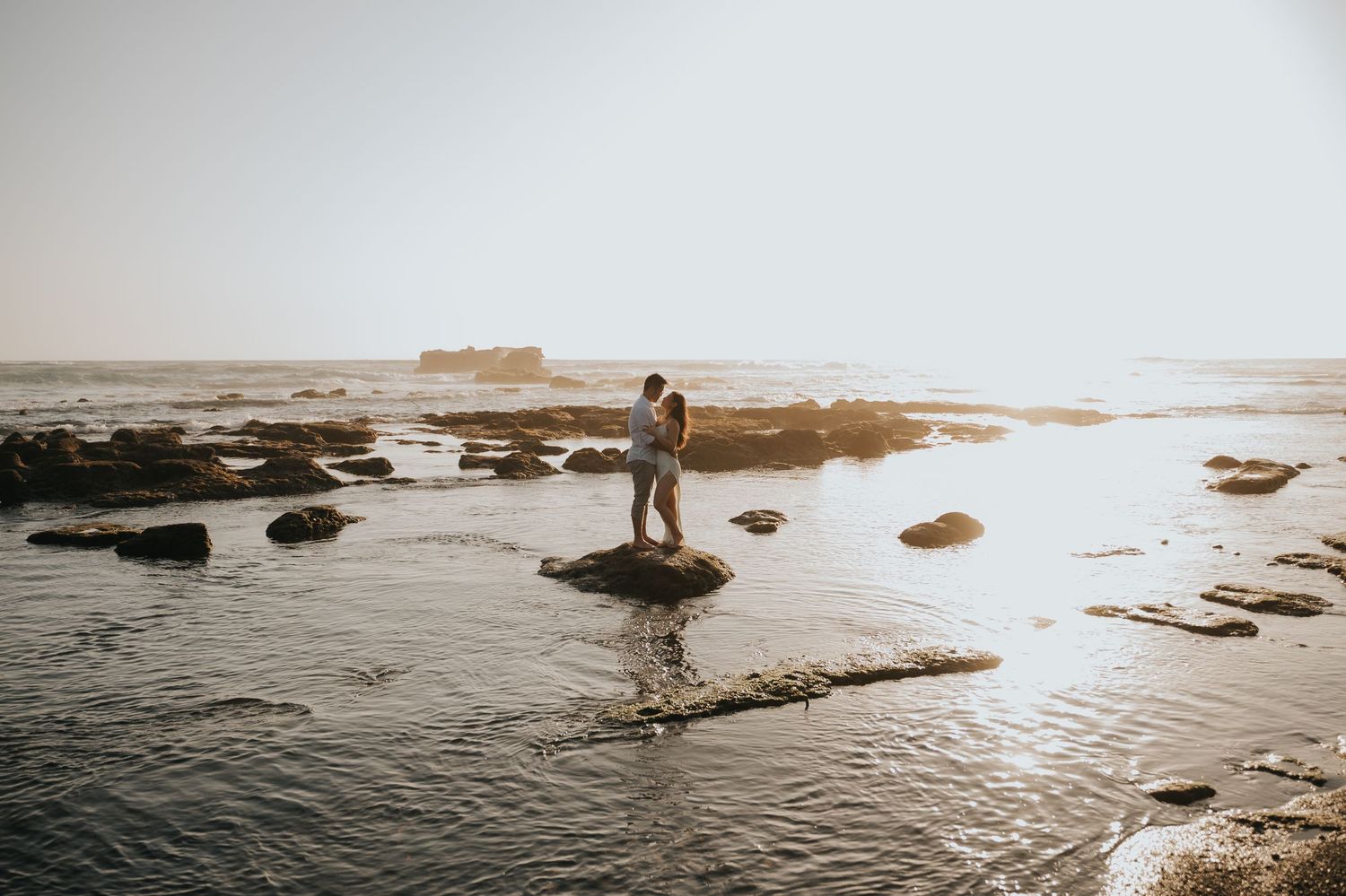 Couple embracing on rocky coastal shore at sunset with calm ocean waters and misty horizon in background.