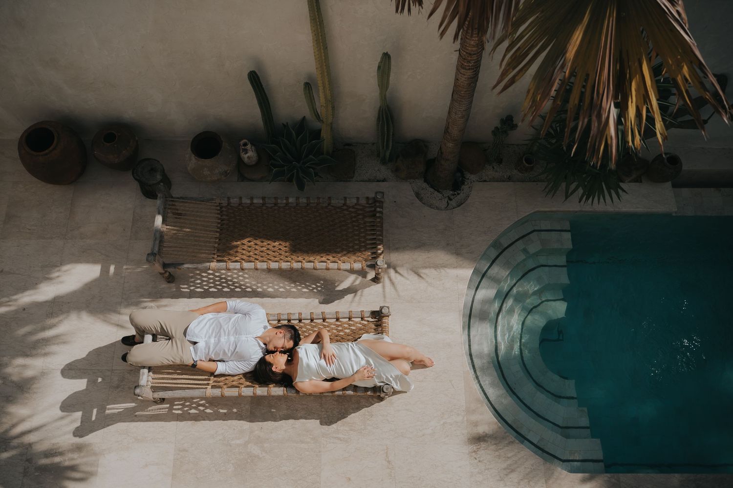 Aerial view of a couple relaxing on a wicker daybed beside a turquoise pool, surrounded by cacti and tropical plants.