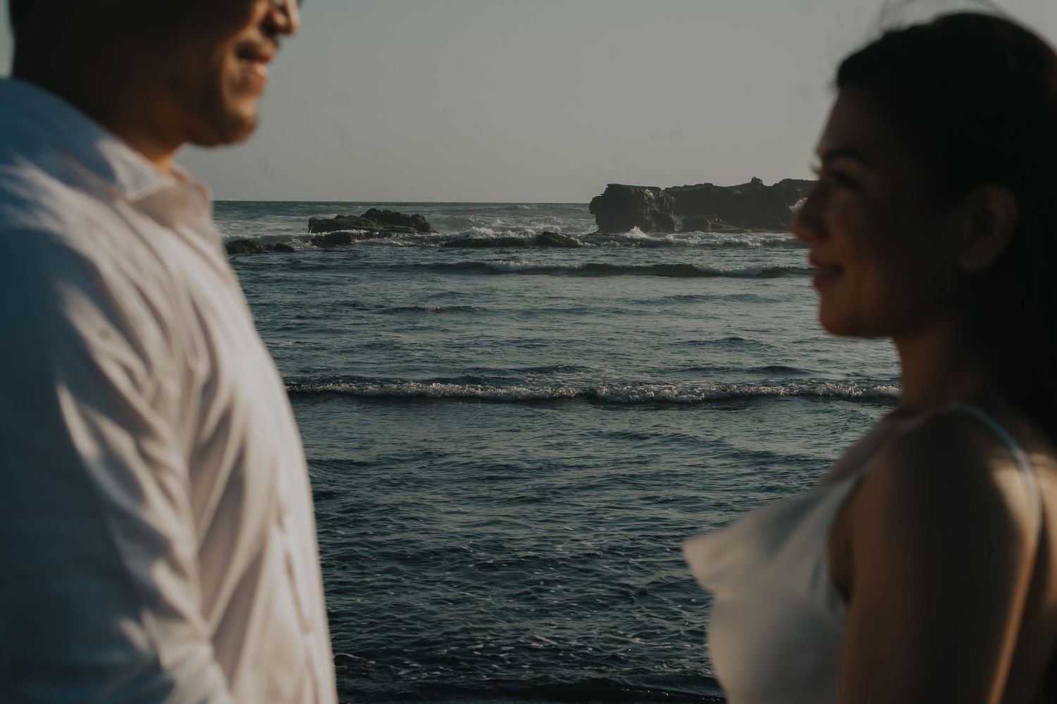 Couple sharing a romantic moment by the ocean at sunset, with gentle waves and rocky coastline in the background.