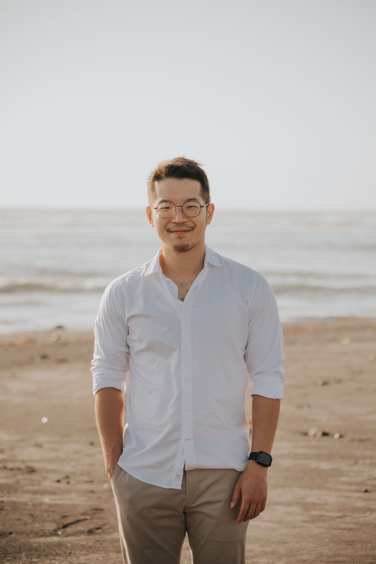 A smiling young Asian man in a white linen shirt and khaki pants stands on a sandy beach with ocean waves behind him.