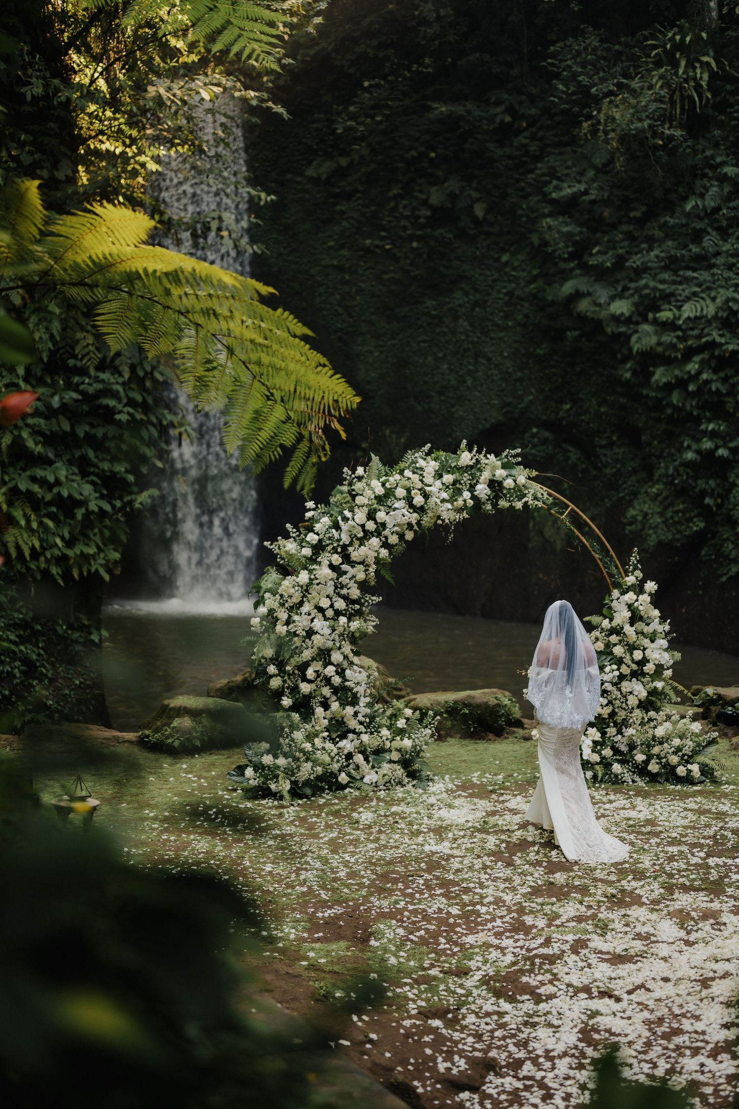 Bride in white gown and veil stands before a circular floral arch near a waterfall in a lush tropical garden.