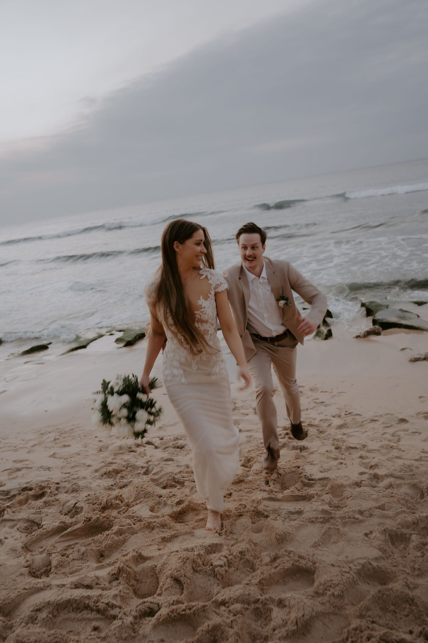 A couple in wedding attire runs playfully along a moody beach shoreline with crashing waves in the background.