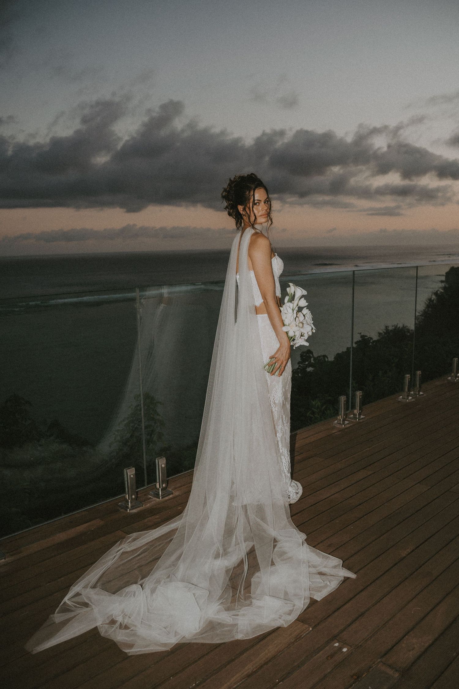A bride in a flowing white gown and veil stands on a wooden deck overlooking a dramatic sunset landscape.