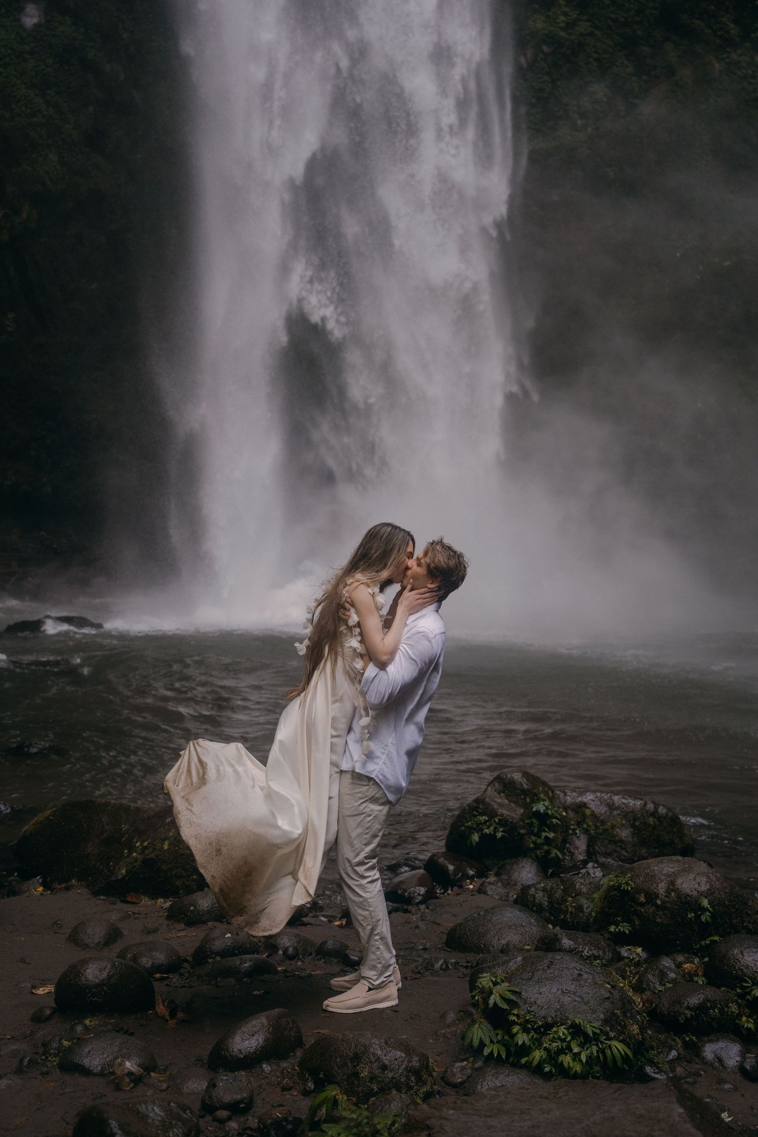 A romantic couple embraces in front of a majestic waterfall, with her dress flowing in the misty breeze.