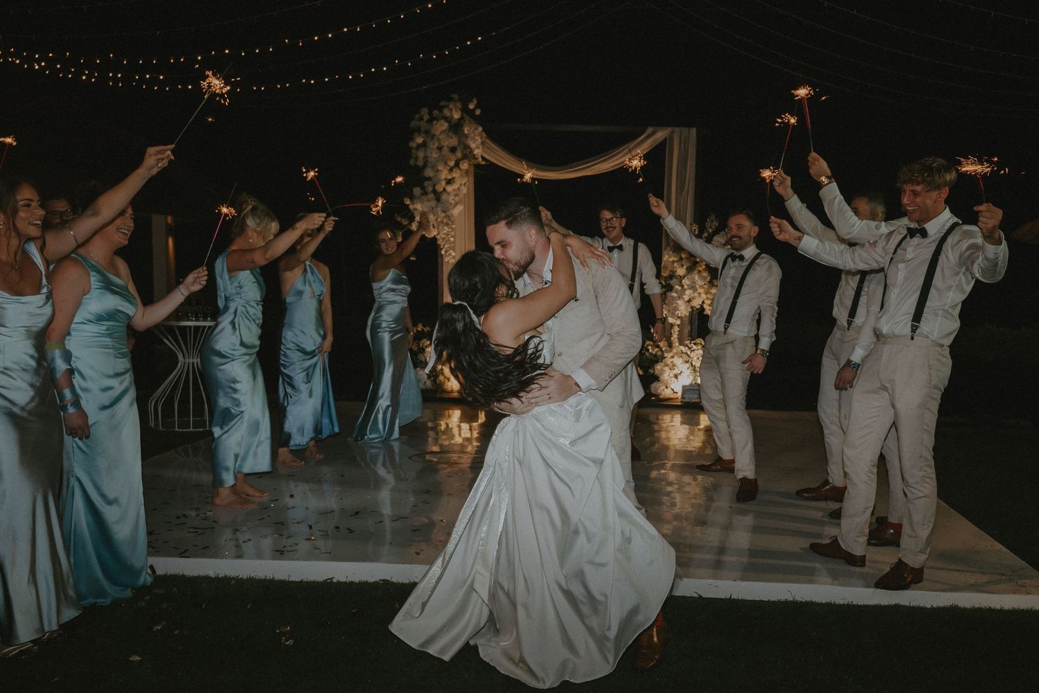 Wedding guests dance together on an outdoor dance floor illuminated by string lights during an evening celebration.
