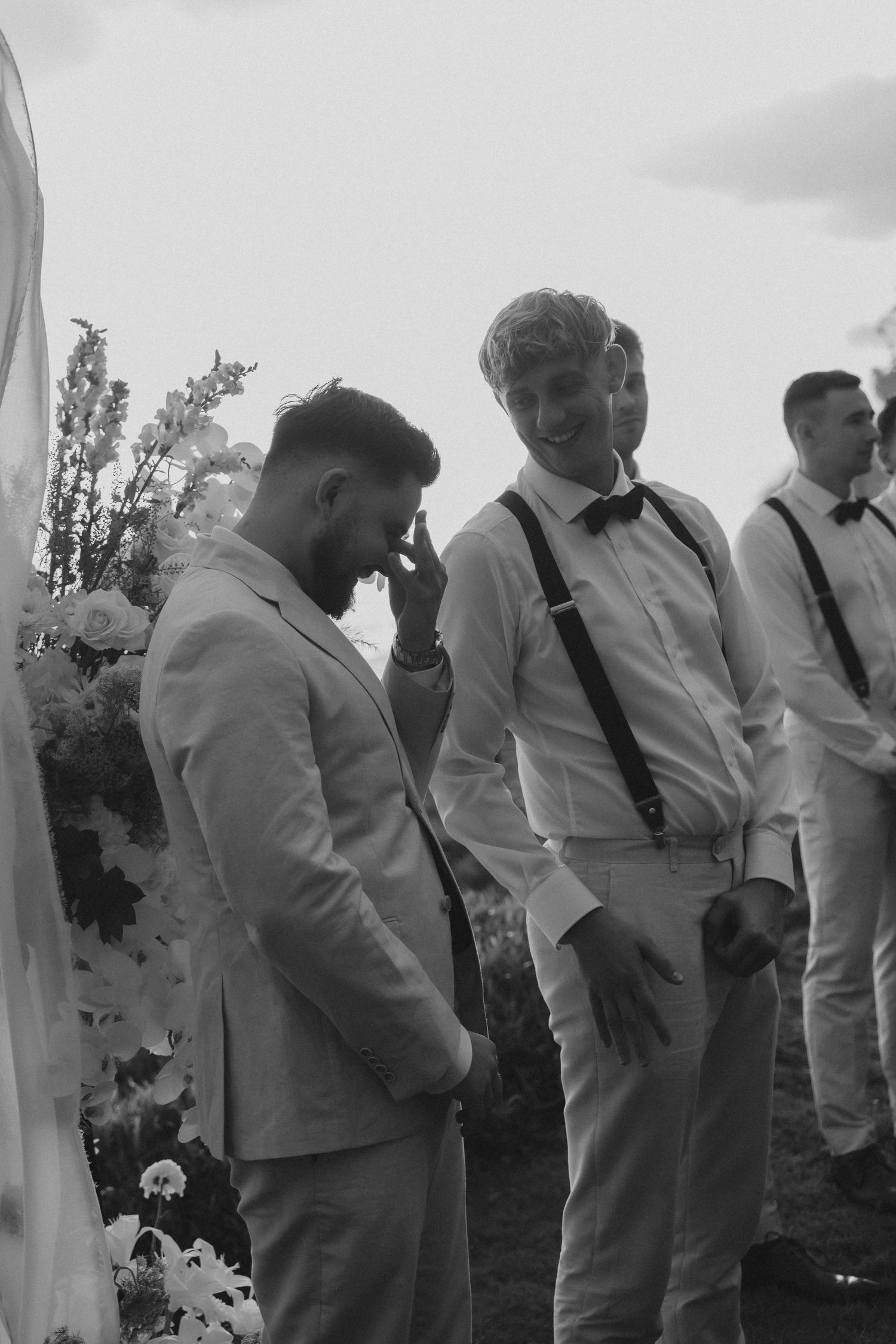 A black and white photo shows groomsmen in light-colored suits standing in a line outdoors at a wedding ceremony.
