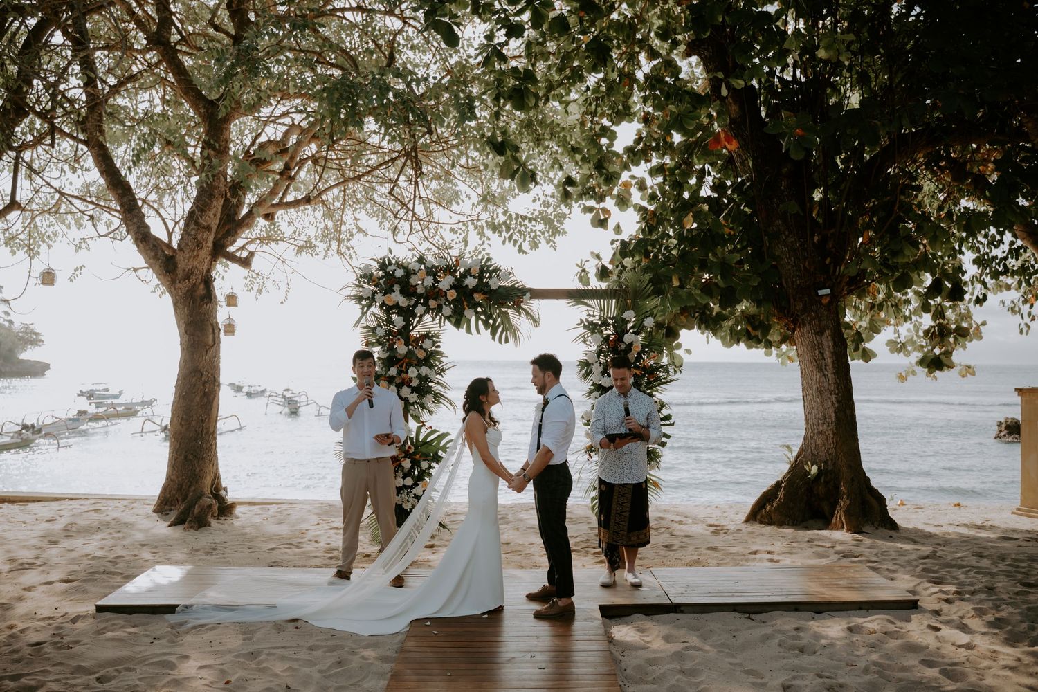 A couple shares an intimate moment under tropical trees on a sandy beach during their wedding ceremony.