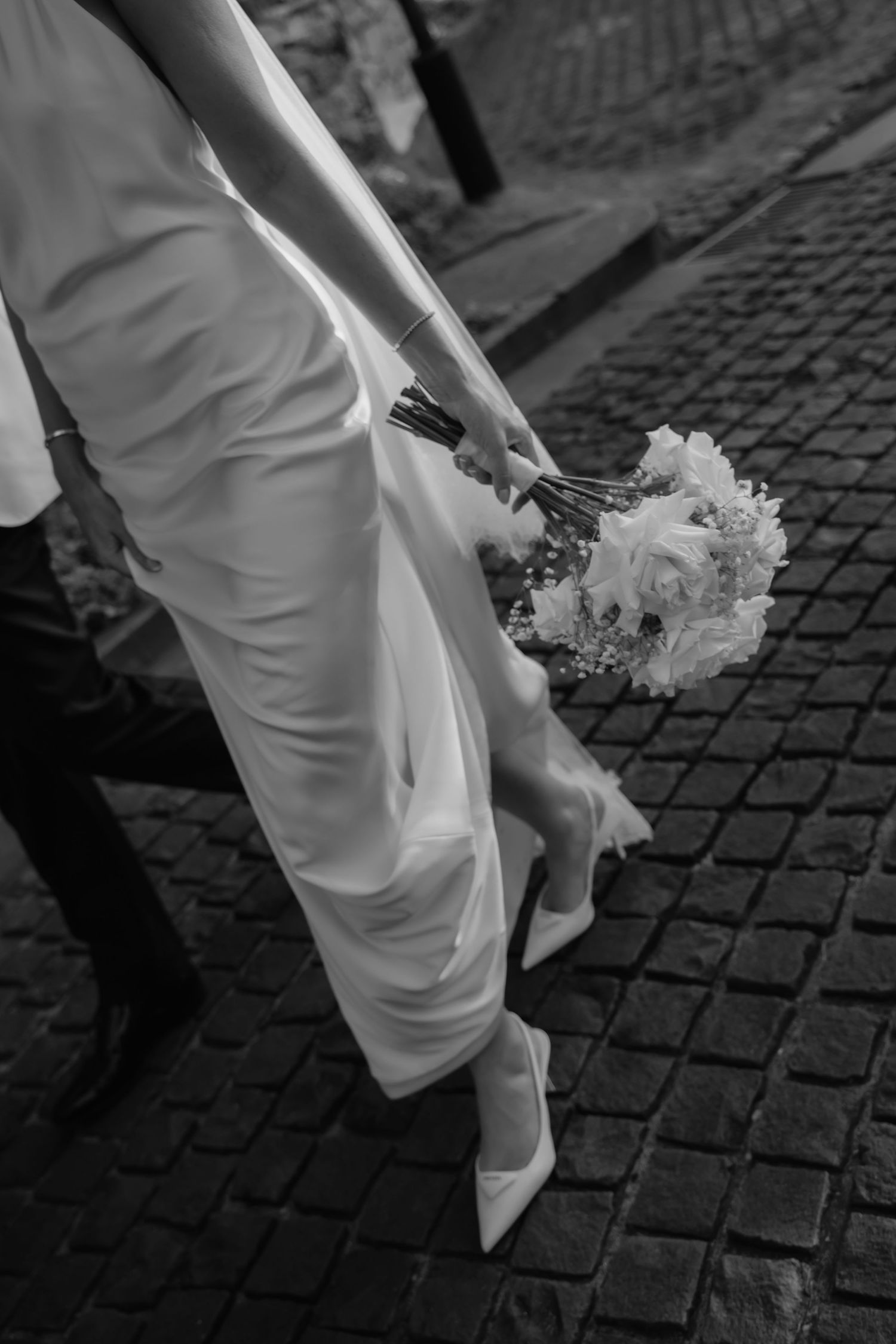 Close-up of bride's white dress and bouquet against cobblestone street in black and white.
