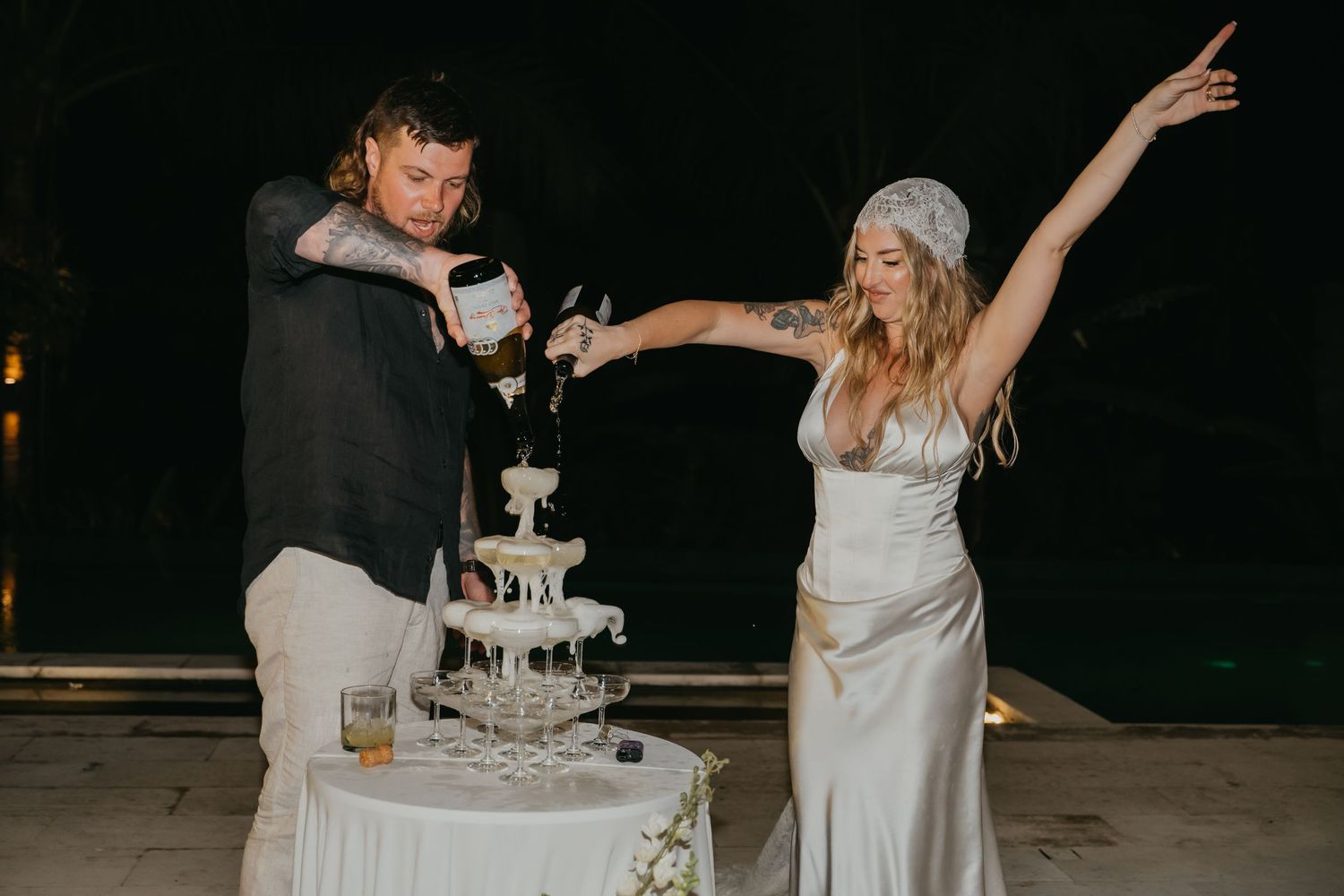 Bride and groom pour champagne tower together at their outdoor wedding reception at night.