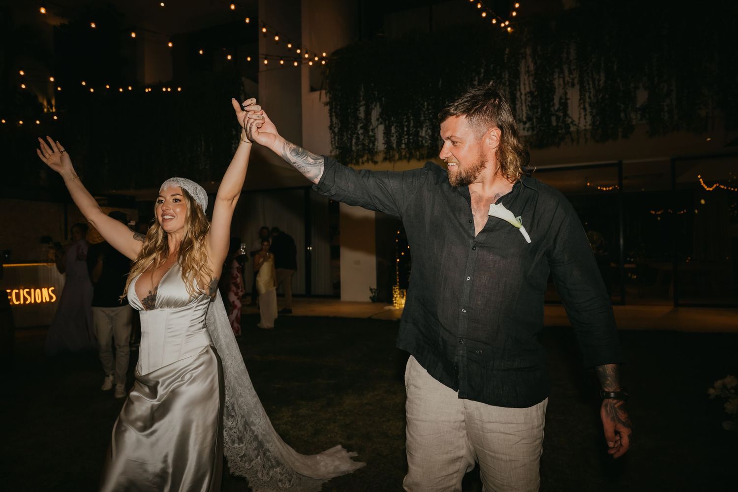 Bride and groom dancing joyfully at outdoor wedding reception under string lights at night.