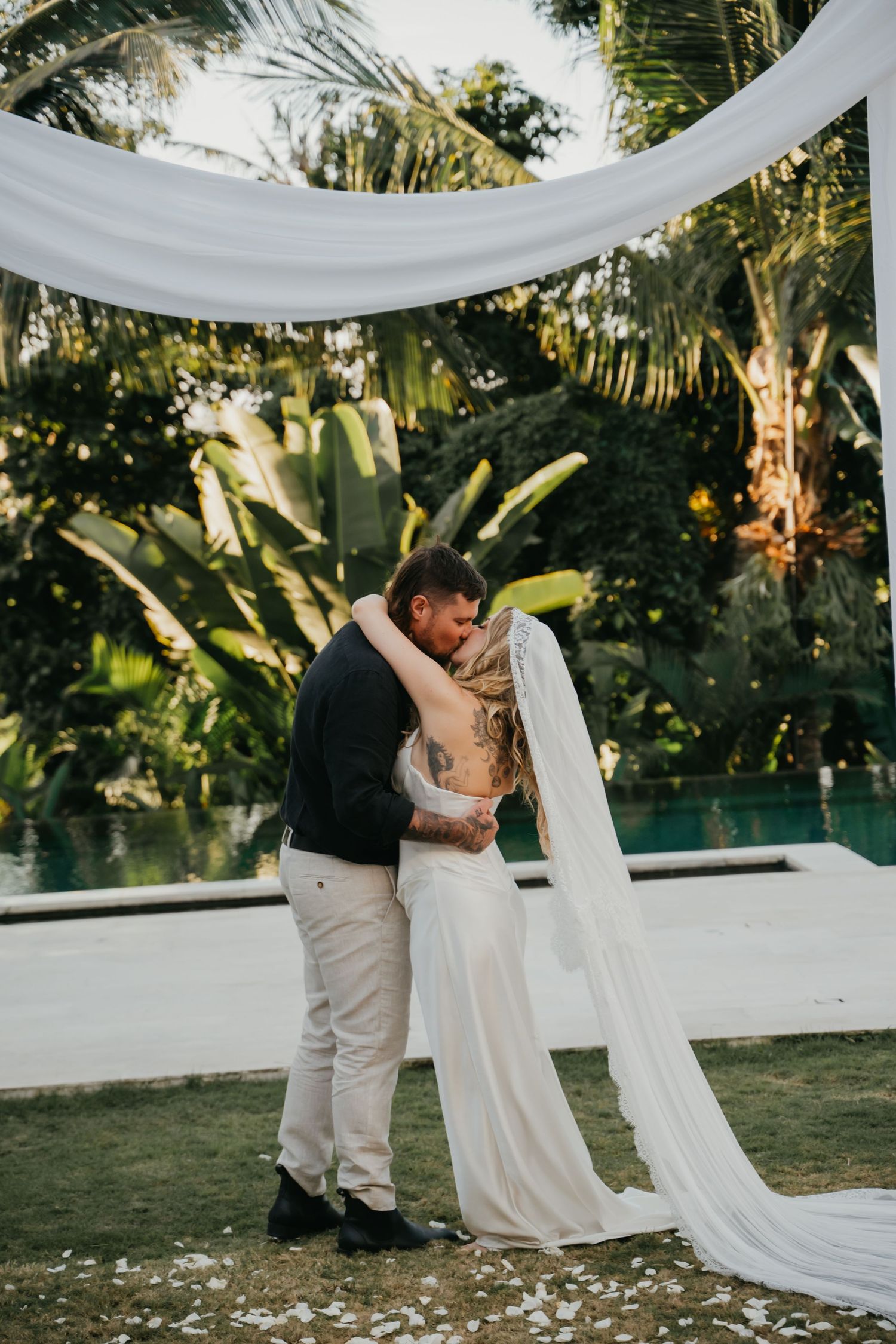 Bride and groom share first kiss under white draped arch surrounded by lush tropical greenery and pool.
