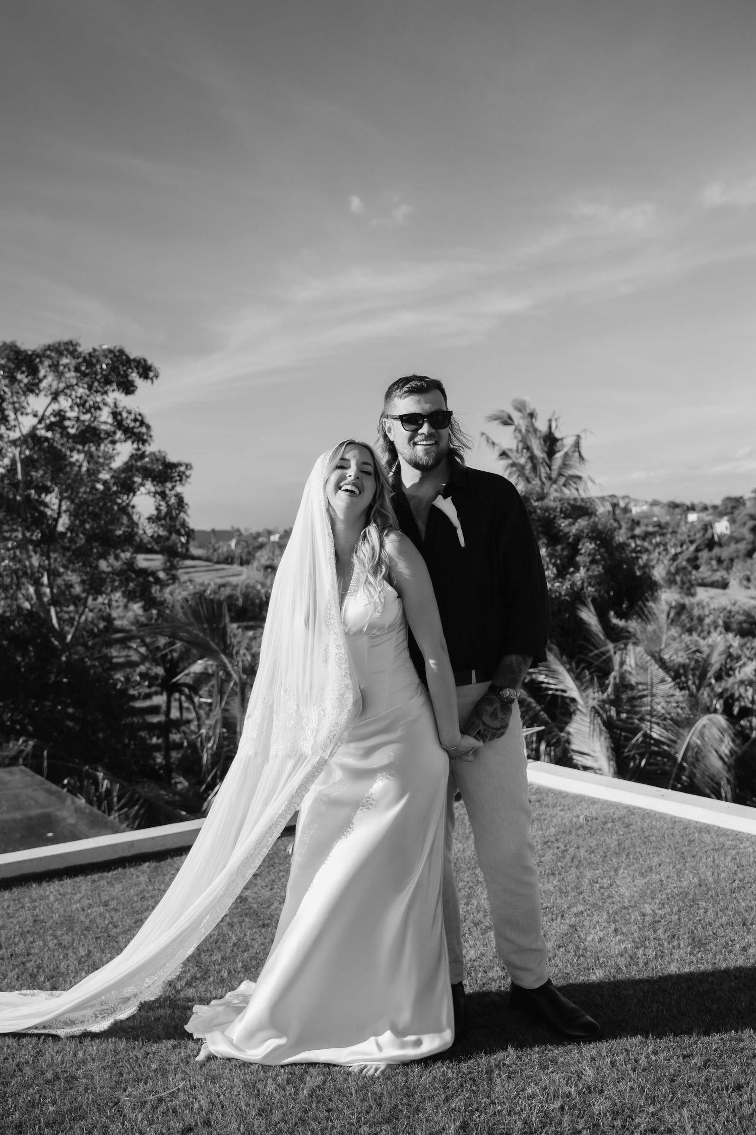 Black and white portrait of smiling bride and groom with veil and palm trees in background.