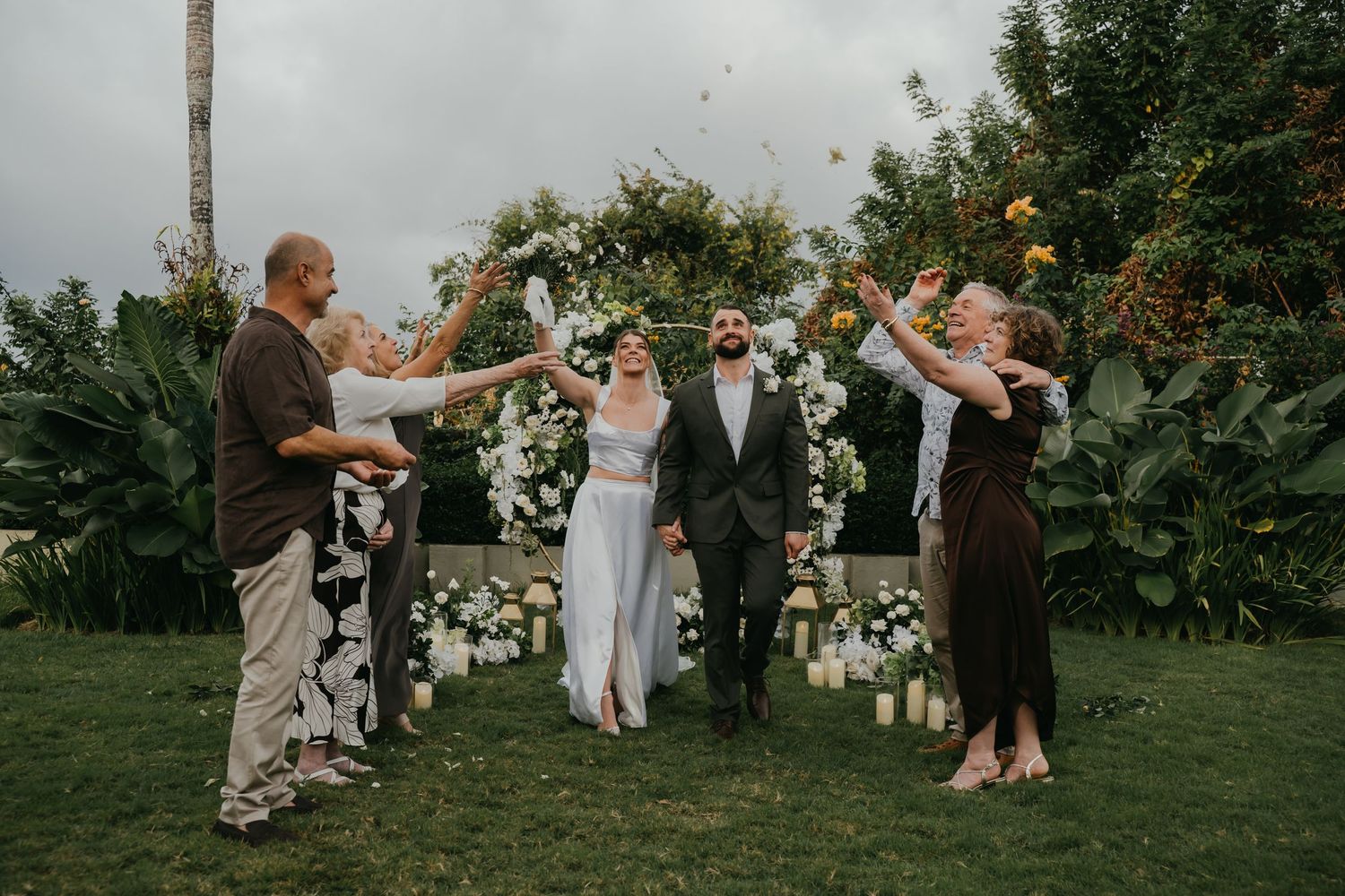 Newlyweds celebrated by guests tossing petals after outdoor garden wedding ceremony.