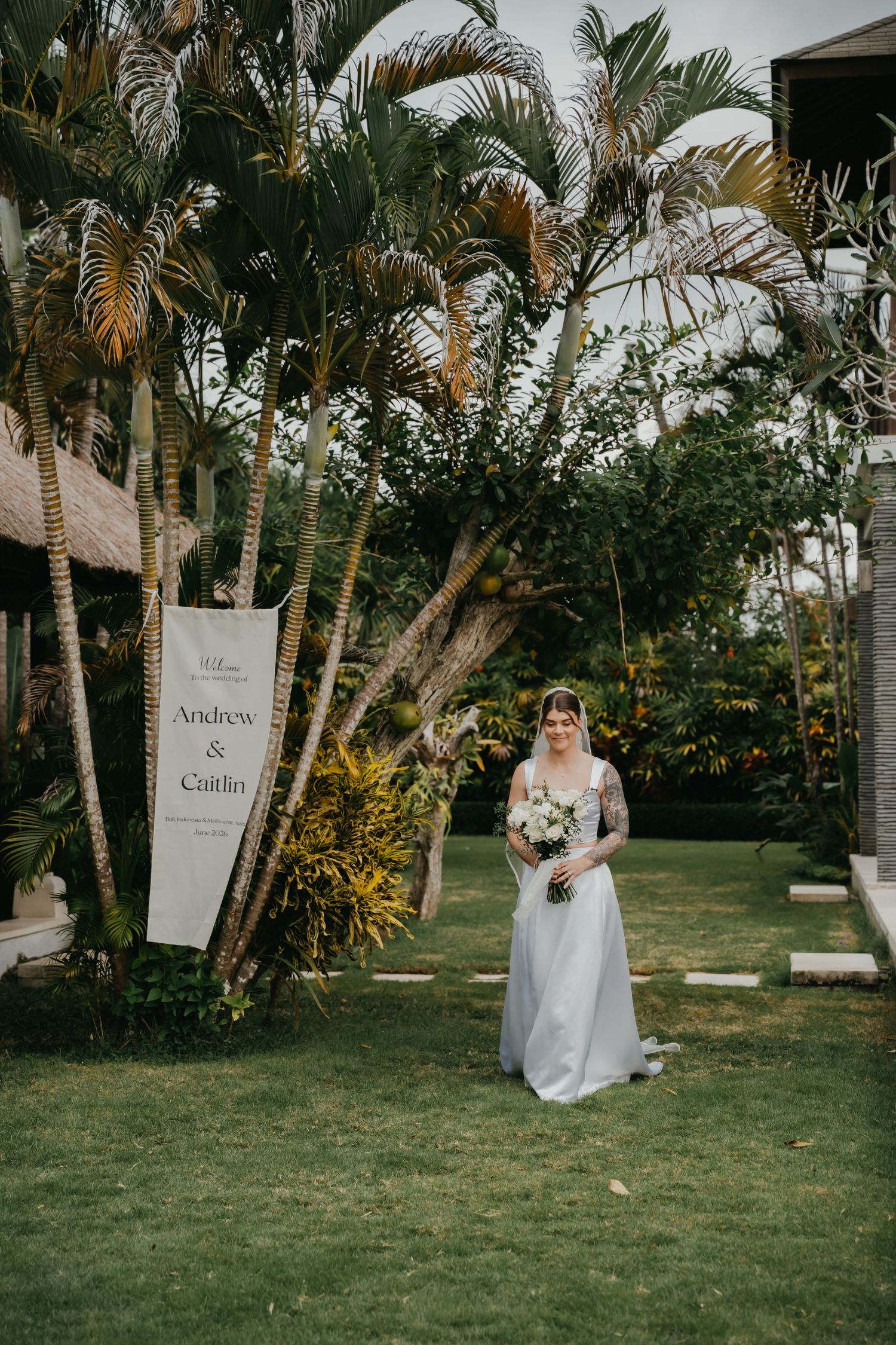 Bride in white gown holding bouquet stands on green lawn beside tropical palm trees and wedding welcome sign.