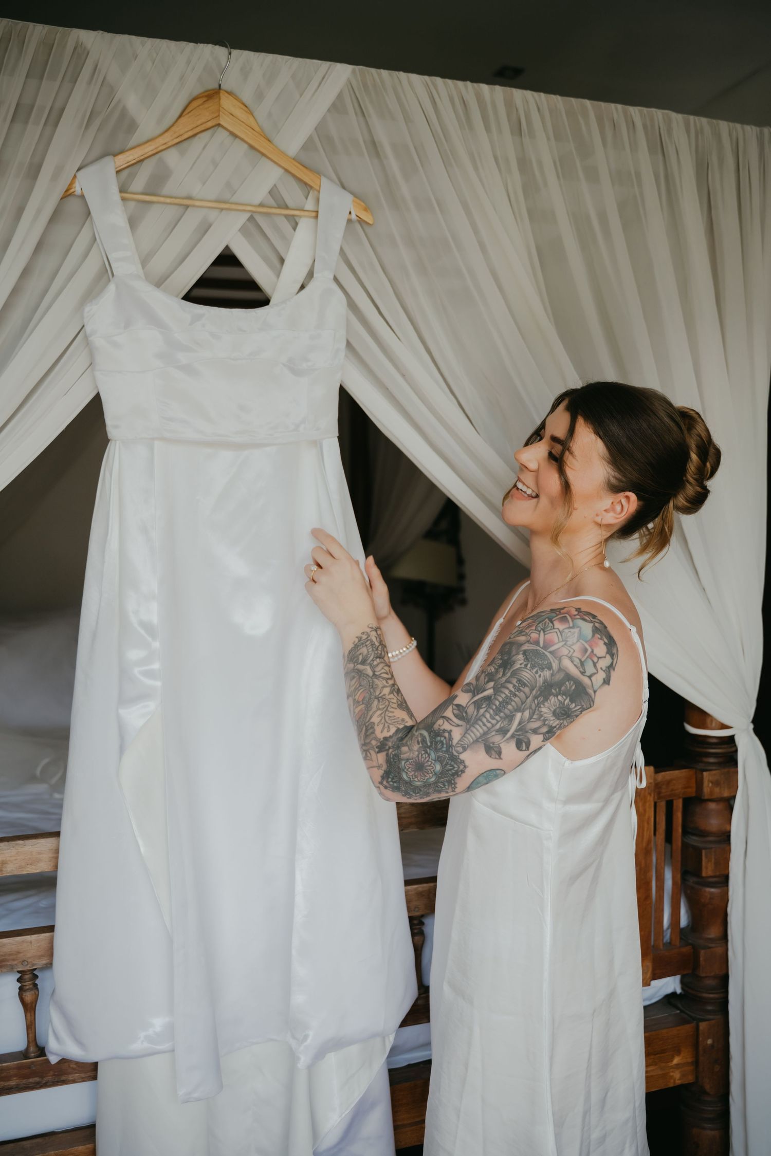 Smiling tattooed bride holds her elegant white wedding dress on a hanger in a romantic canopy bedroom.