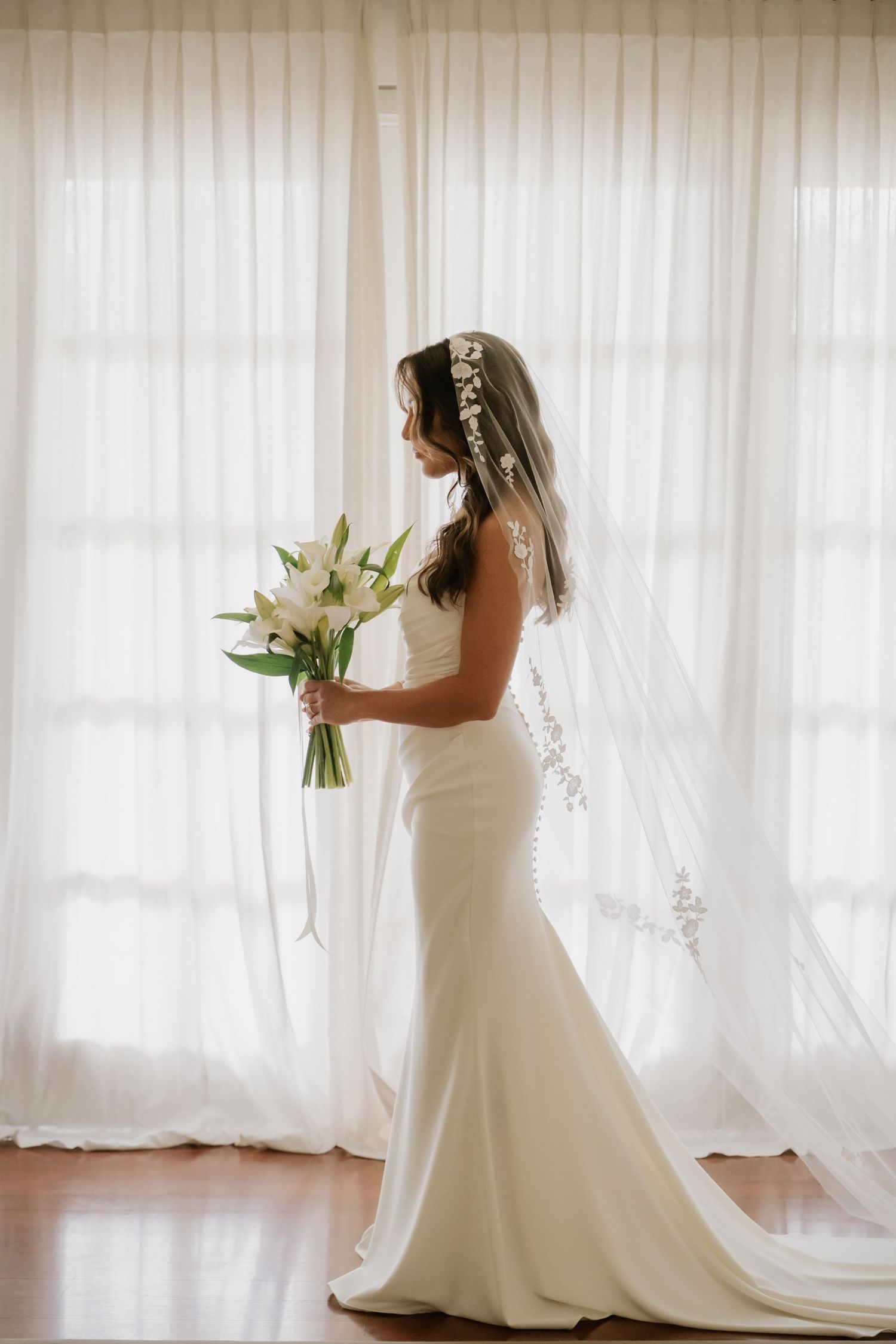 Elegant bride in fitted white gown with lace veil holding white lily bouquet by sheer curtains.