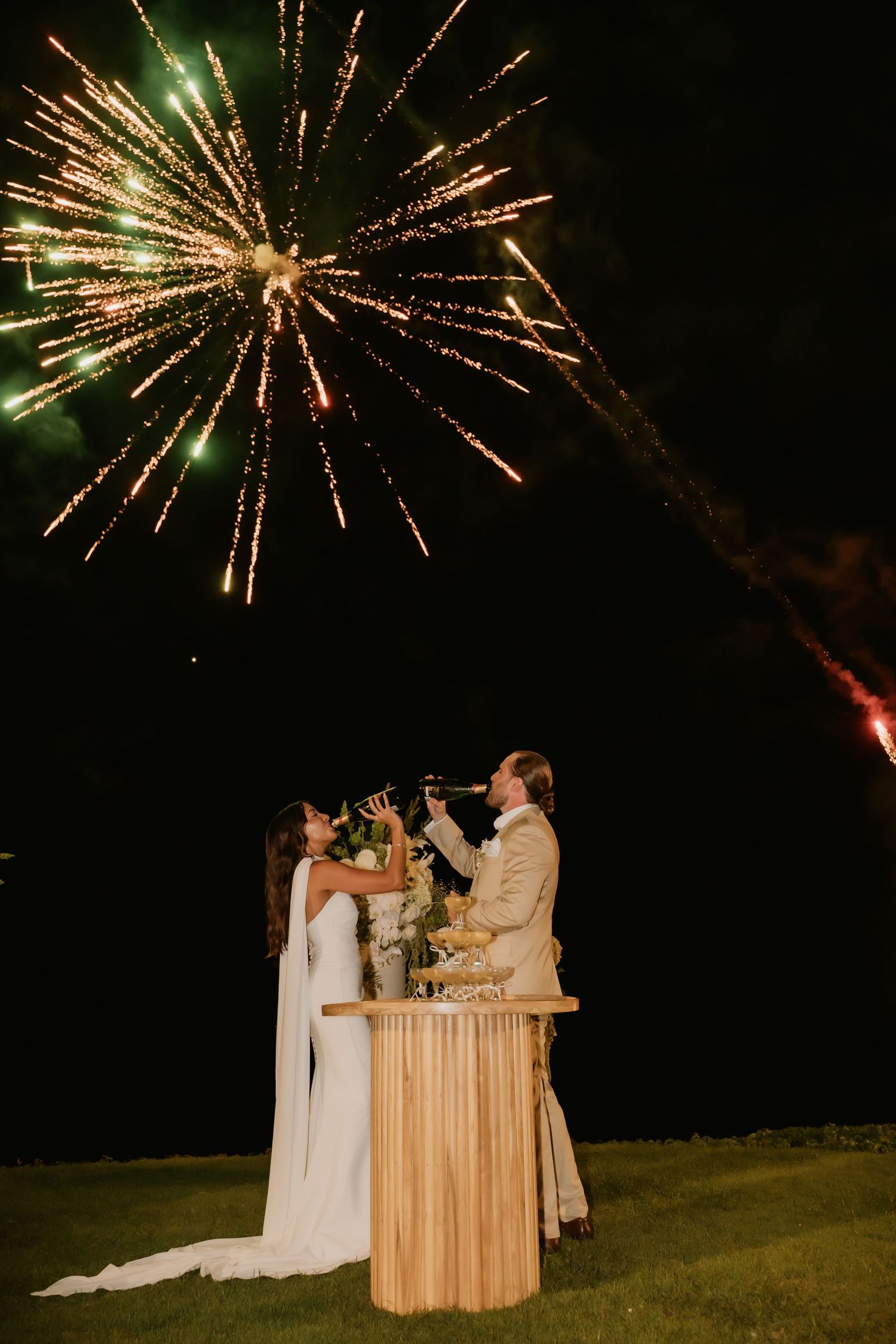 Bride and groom toast champagne beneath spectacular fireworks display at nighttime wedding celebration.