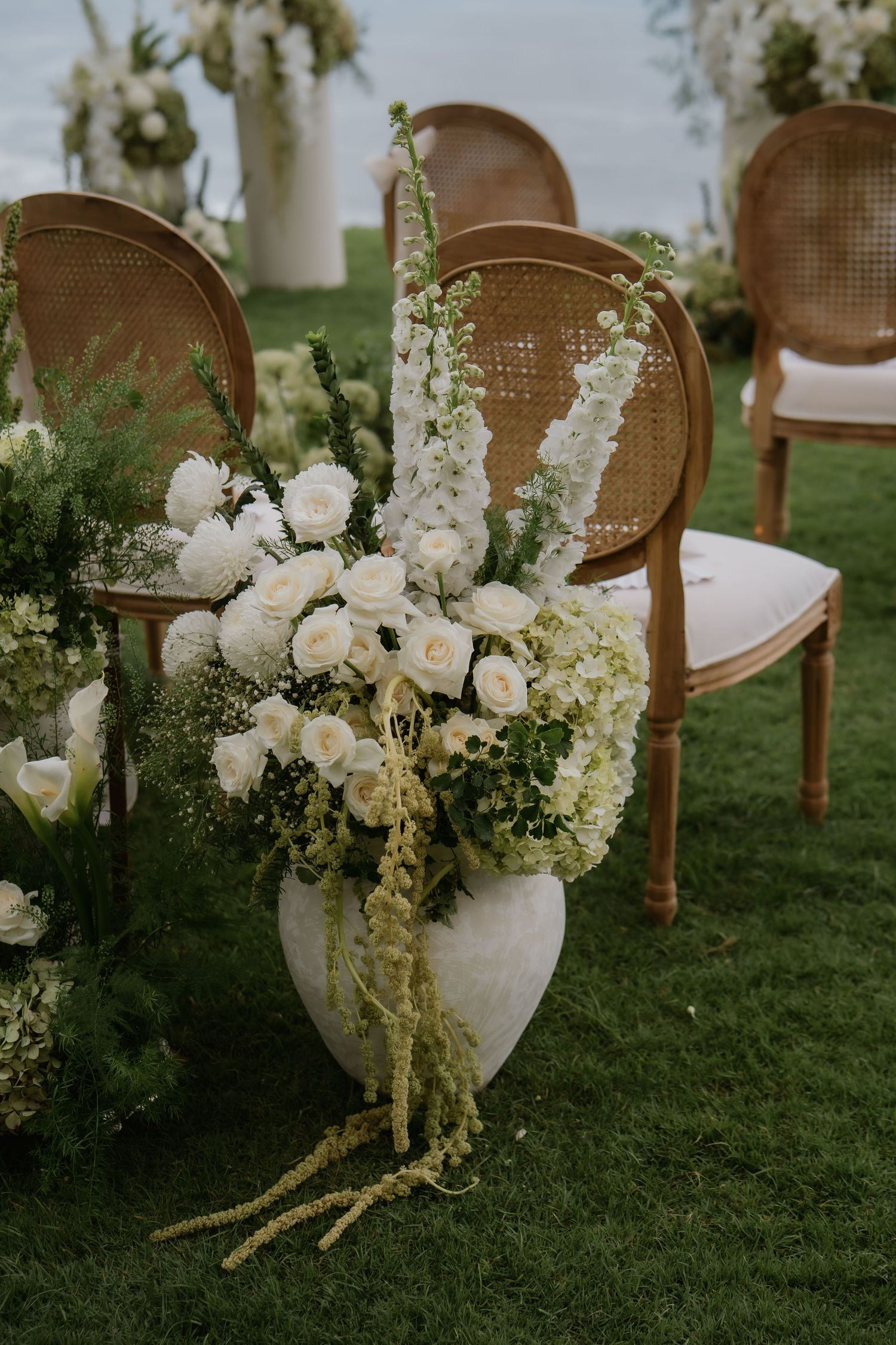 White roses and hydrangeas in a round stone vase line the aisle beside rattan chairs at an outdoor wedding.