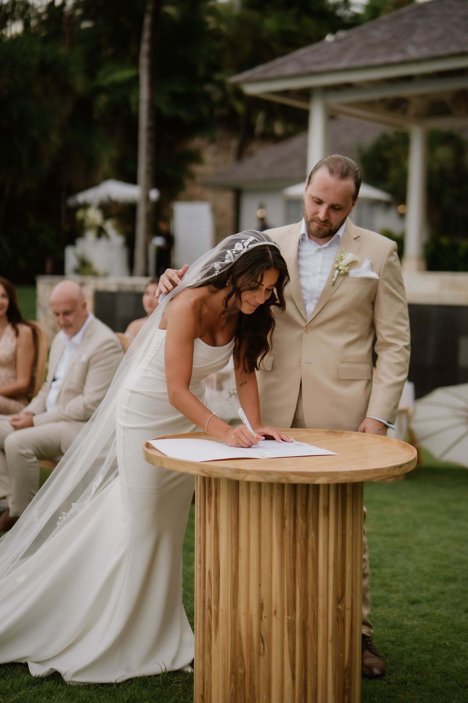 Bride signs wedding certificate at a wooden table while groom watches during outdoor ceremony.