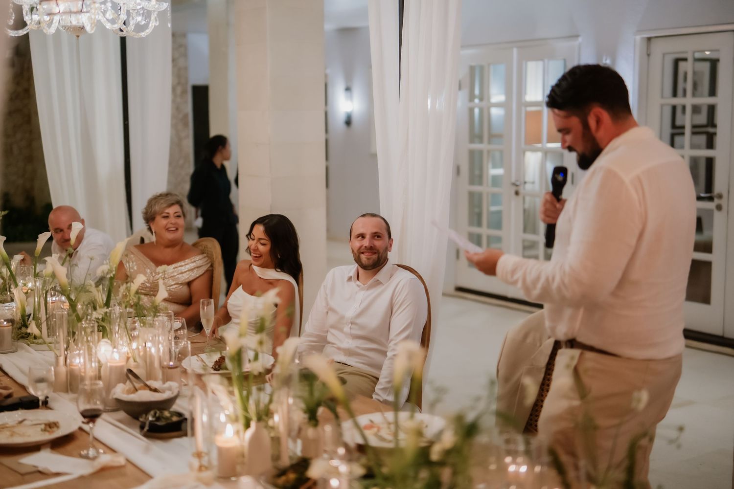 Best man gives toast at wedding reception as bride and groom smile at table.