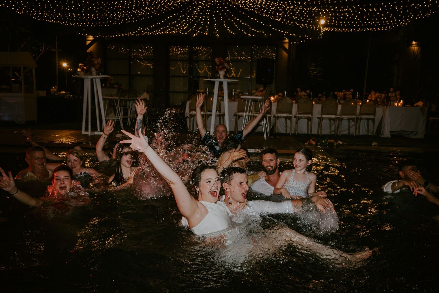 Wedding guests celebrating in pool at night under string lights with joyful splashing and cheering.