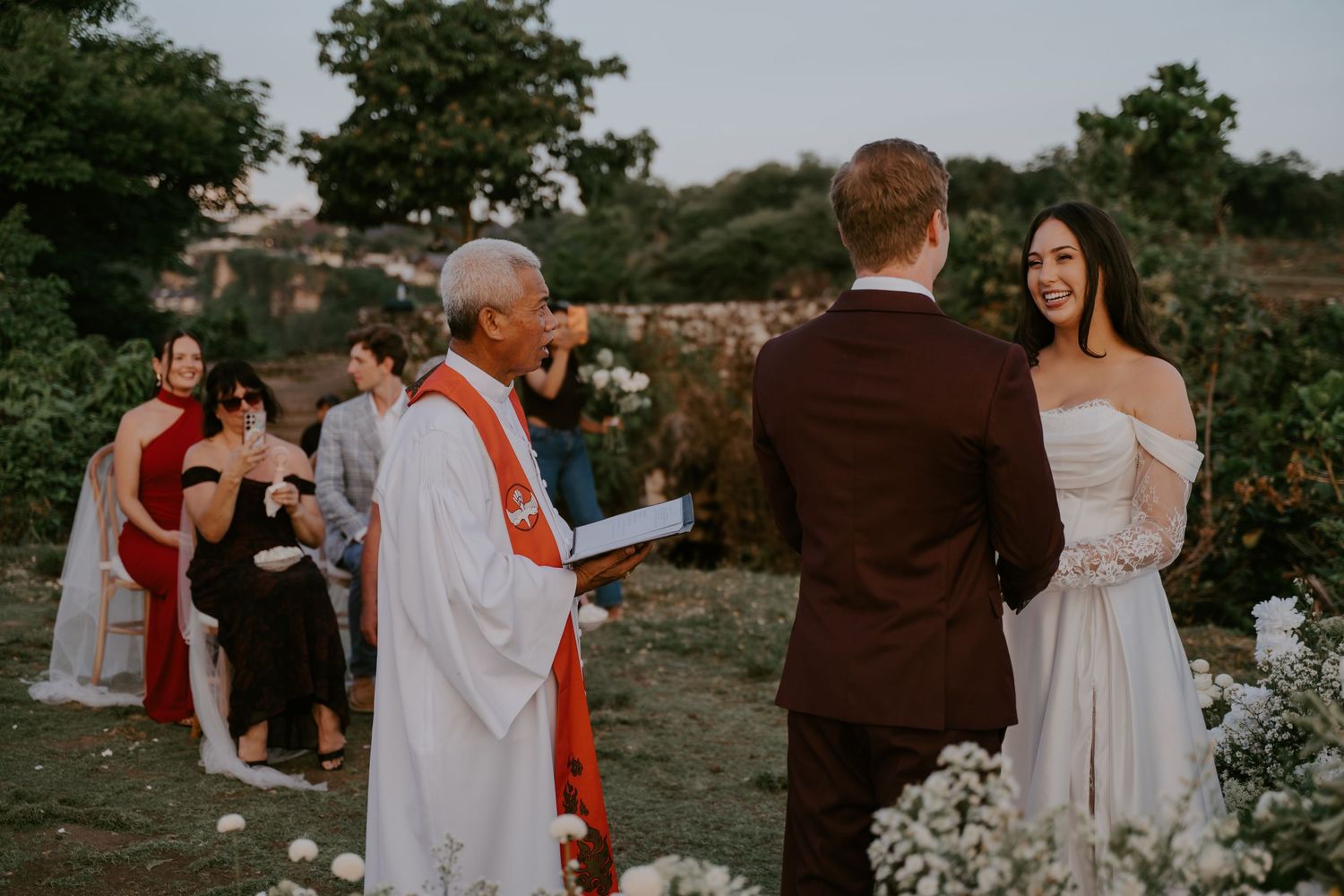 Officiant laughs while reading ceremony vows as bride smiles at groom during outdoor wedding ceremony.