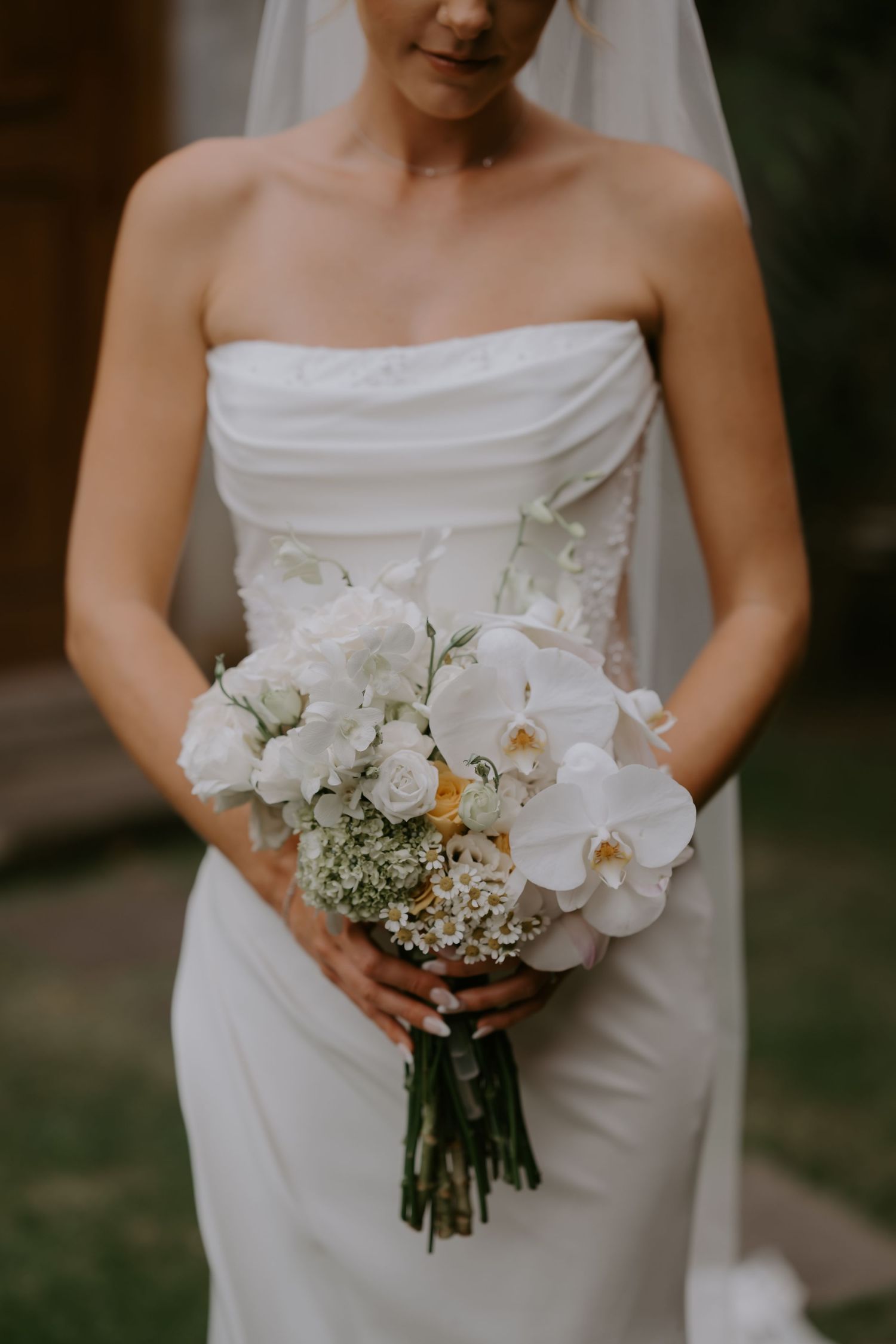 Bride in strapless white gown holding elegant bouquet of white orchids and roses with veil.