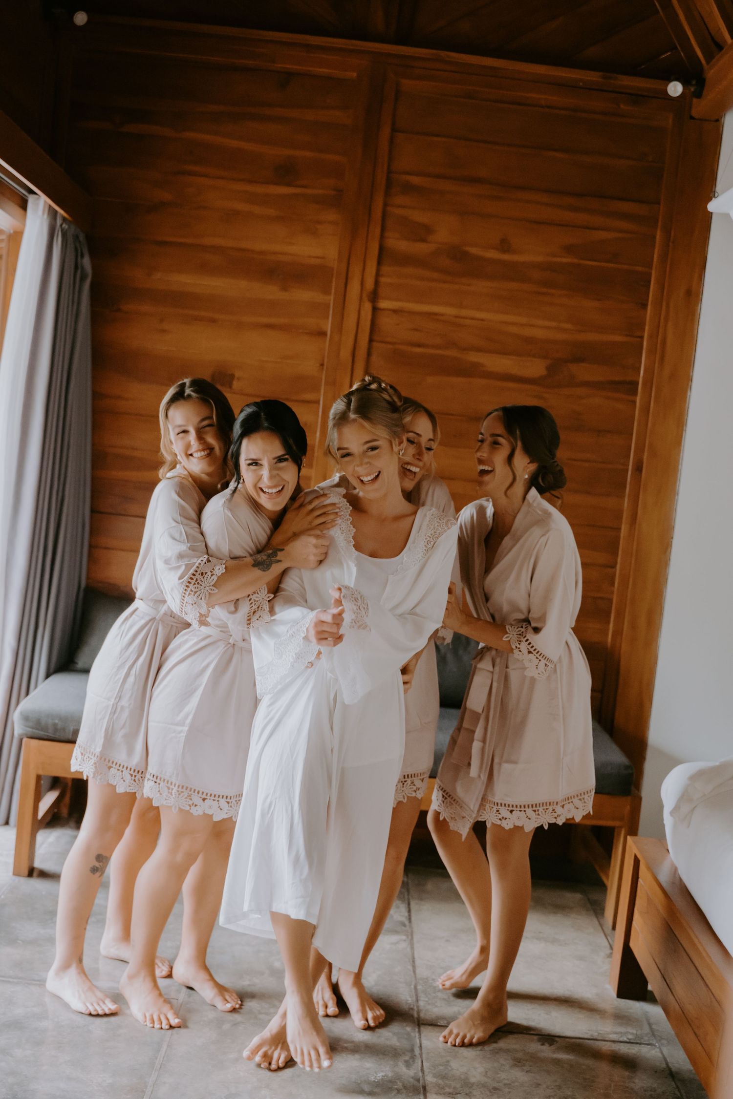 Bride in white robe laughing with three bridesmaids in matching robes inside a wooden cabin room.