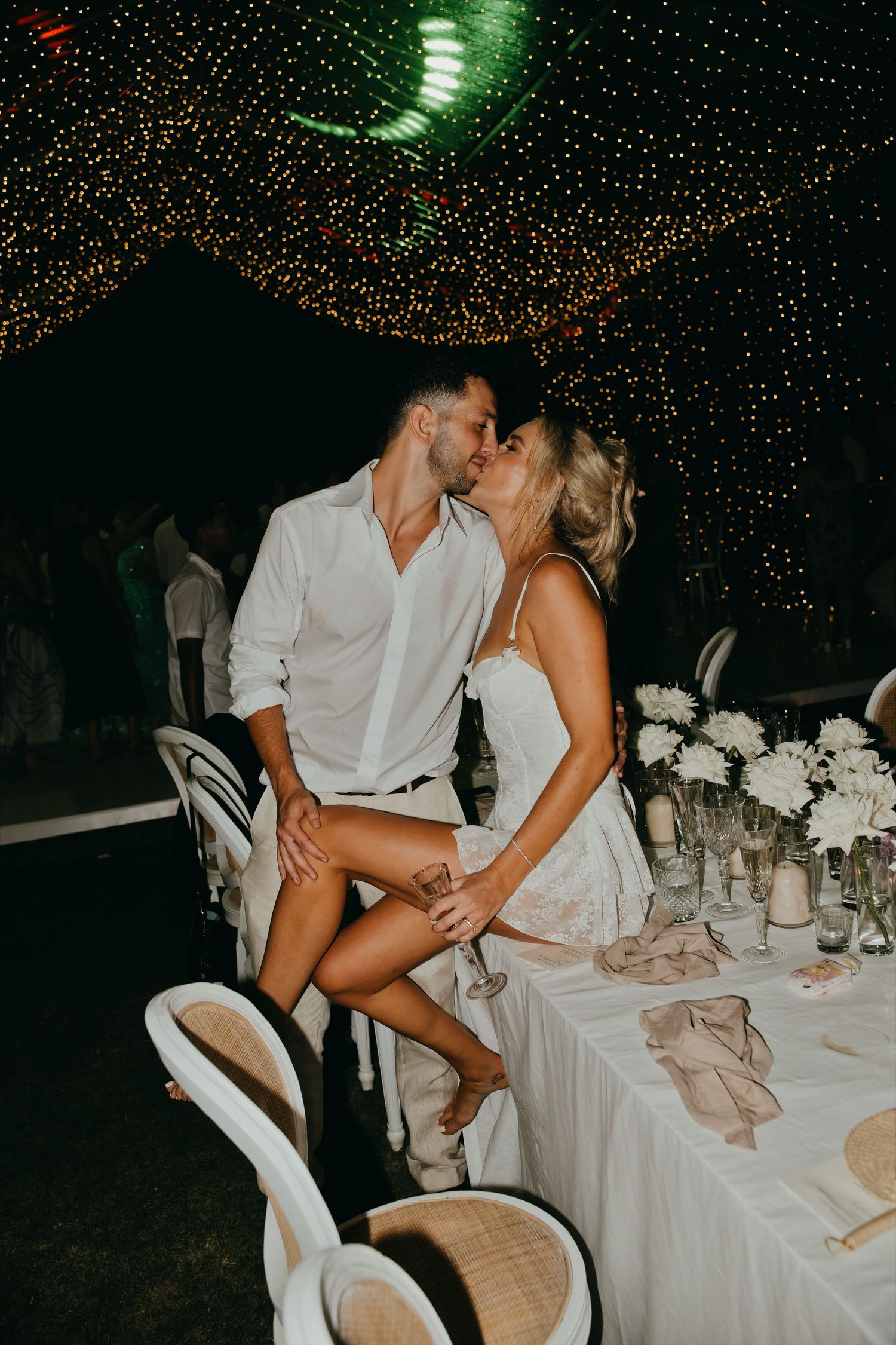 Bride and groom sharing a romantic kiss at the reception table under disco ball and fairy lights.