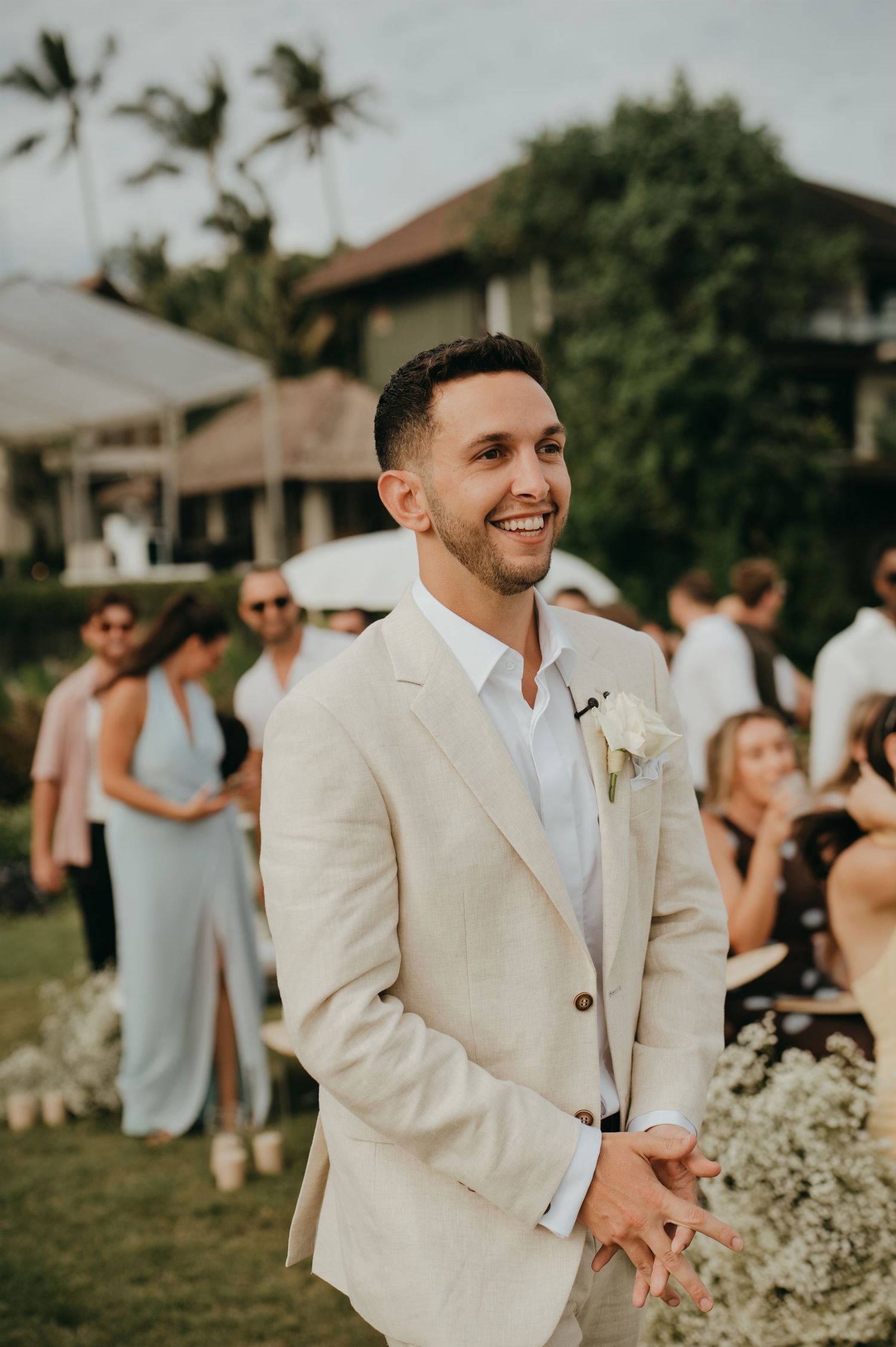 Smiling groom in beige linen suit with white boutonniere at outdoor tropical wedding ceremony.