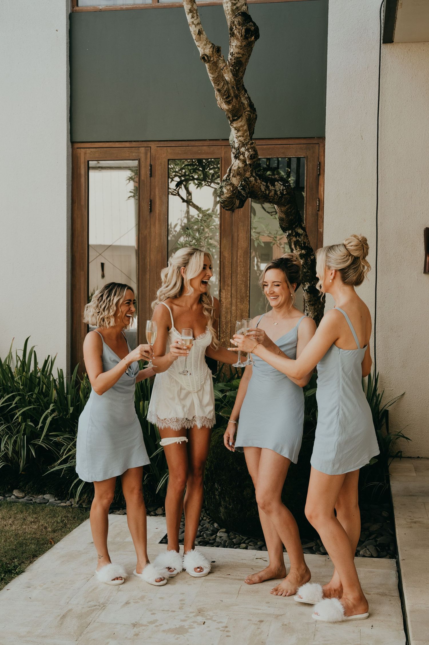 Bride and bridesmaids in matching grey slip dresses toast champagne outside a tropical villa.