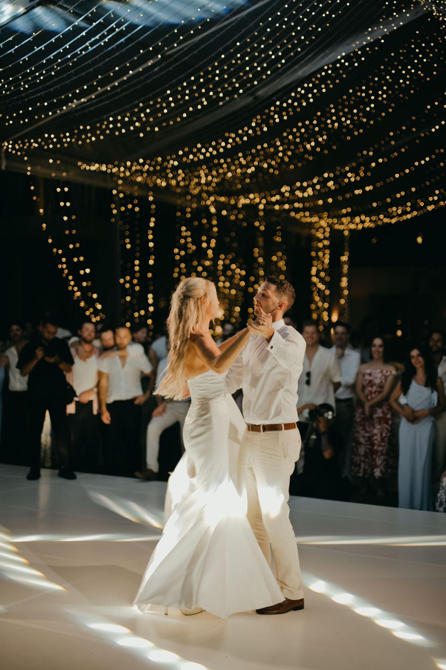 Bride and groom share their first dance under warm string lights while wedding guests watch in background.