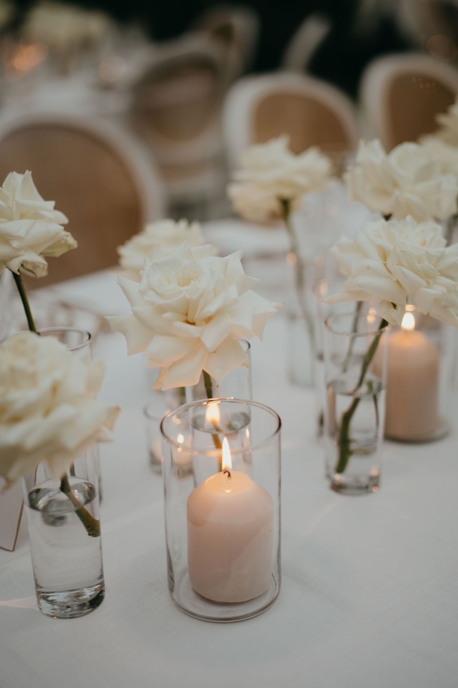 Elegant wedding reception table with white roses in glass vases and lit pillar candles.