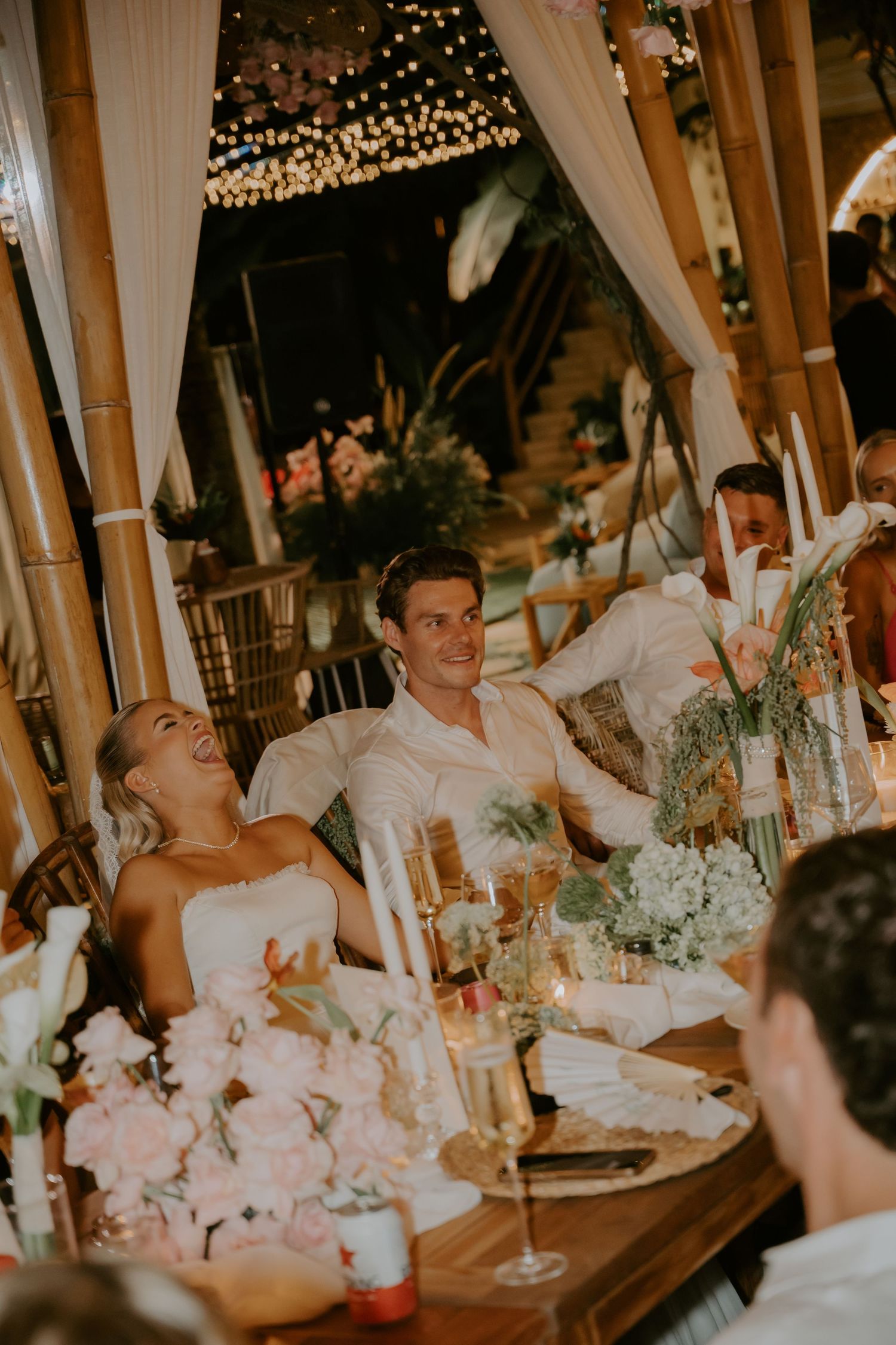Bride and groom laughing joyfully at sweetheart table during outdoor tropical wedding reception at night.