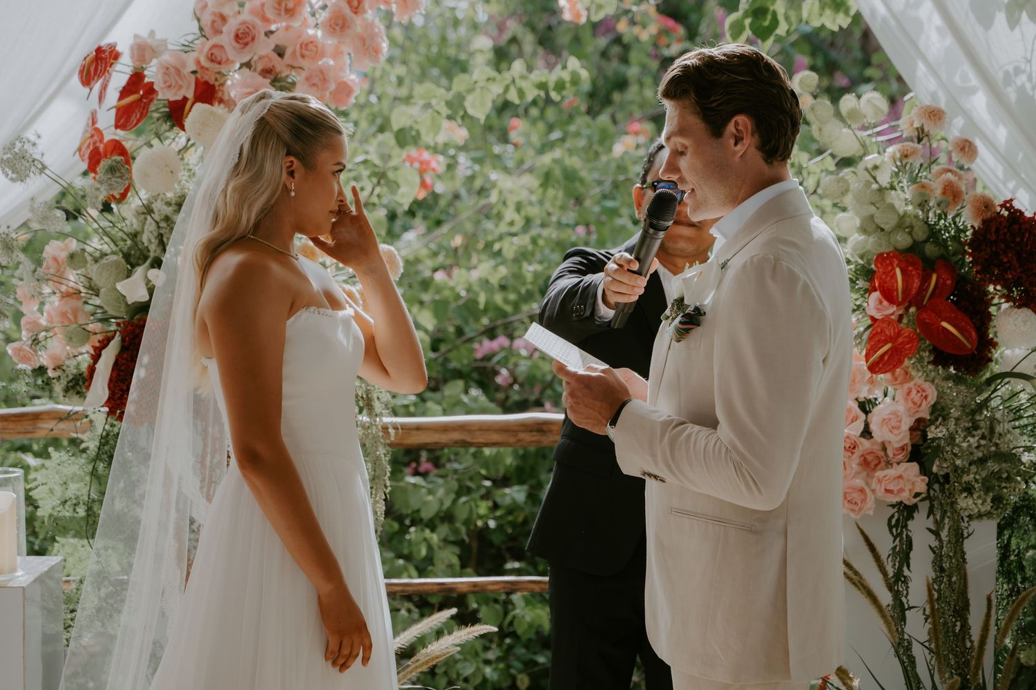 Emotional groom reads wedding vows to tearful bride at outdoor ceremony with floral arch background.