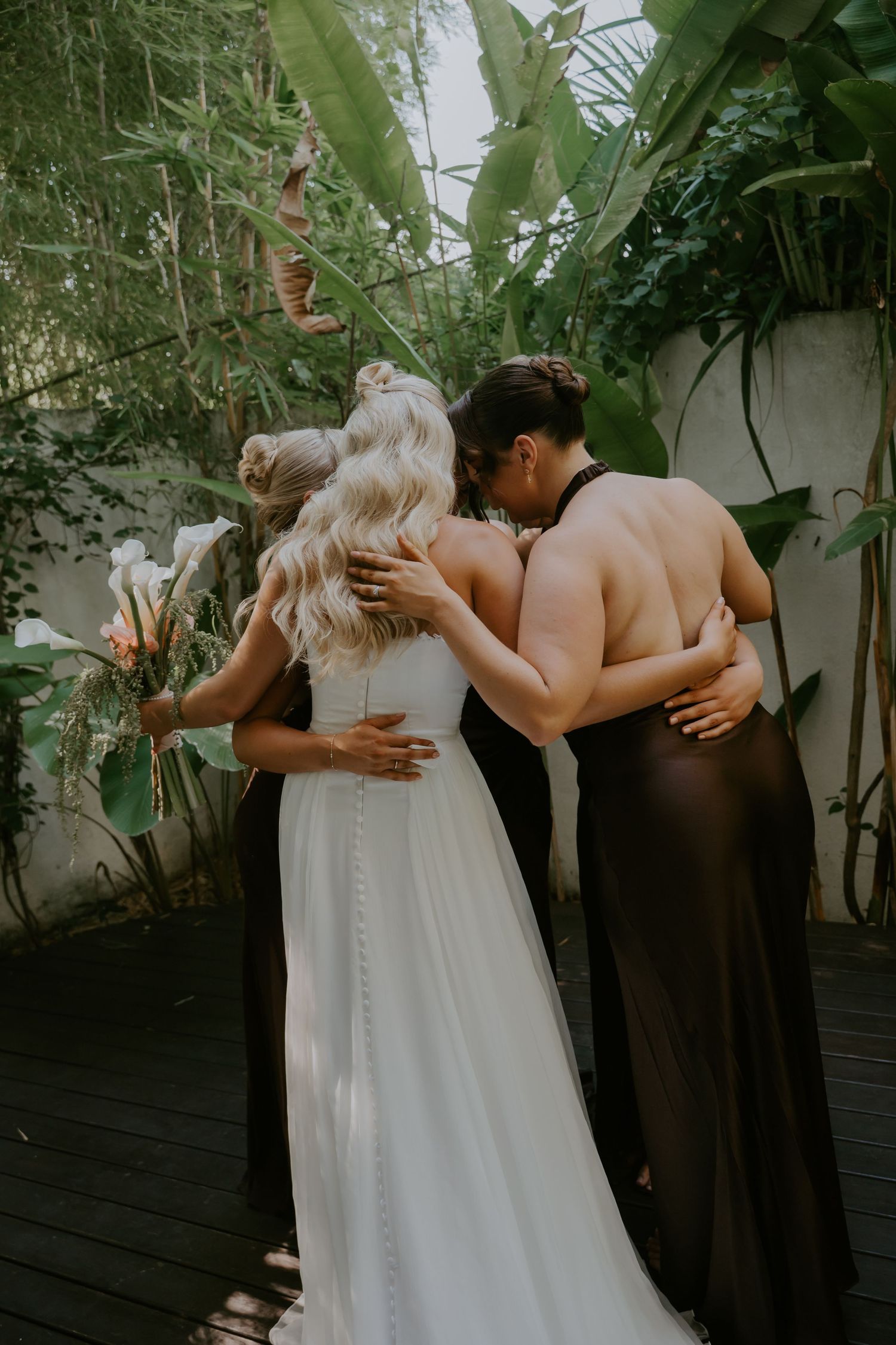 Bride in white gown embracing bridesmaids in lush tropical garden setting during wedding celebration.