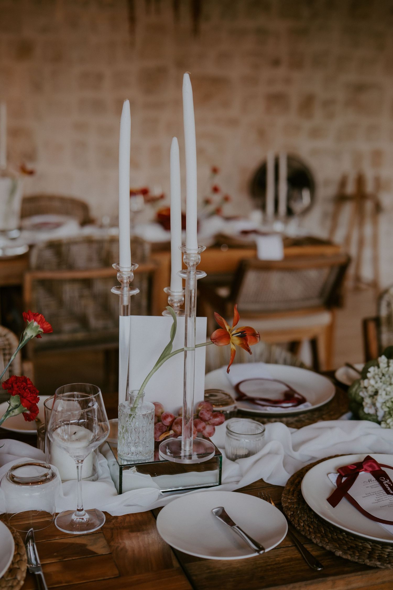 Elegant wedding table setting with tall white taper candles, crystal glasses, and red floral accents.