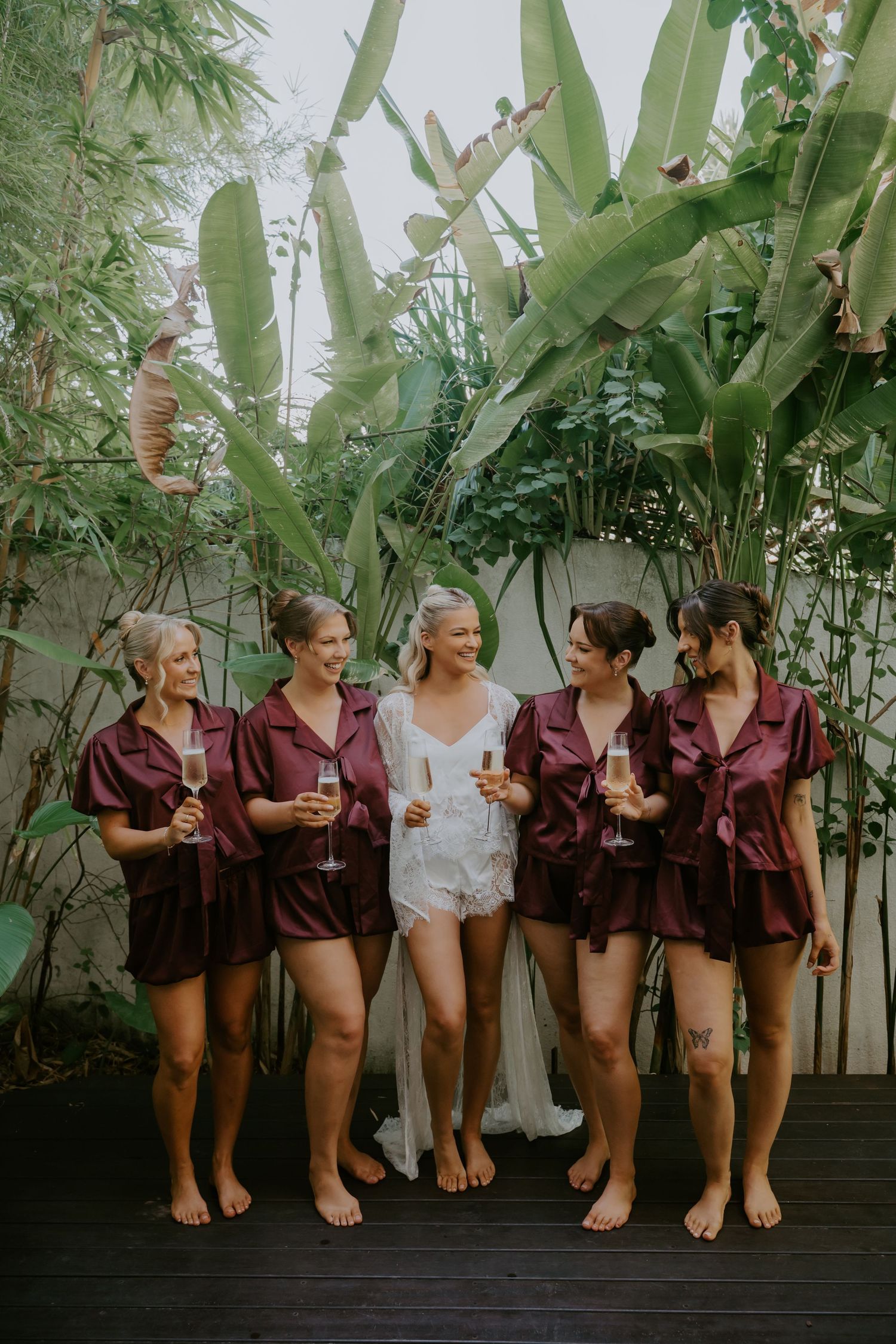 Bride in white lace romper poses with bridesmaids in burgundy satin sets holding champagne among tropical greenery.