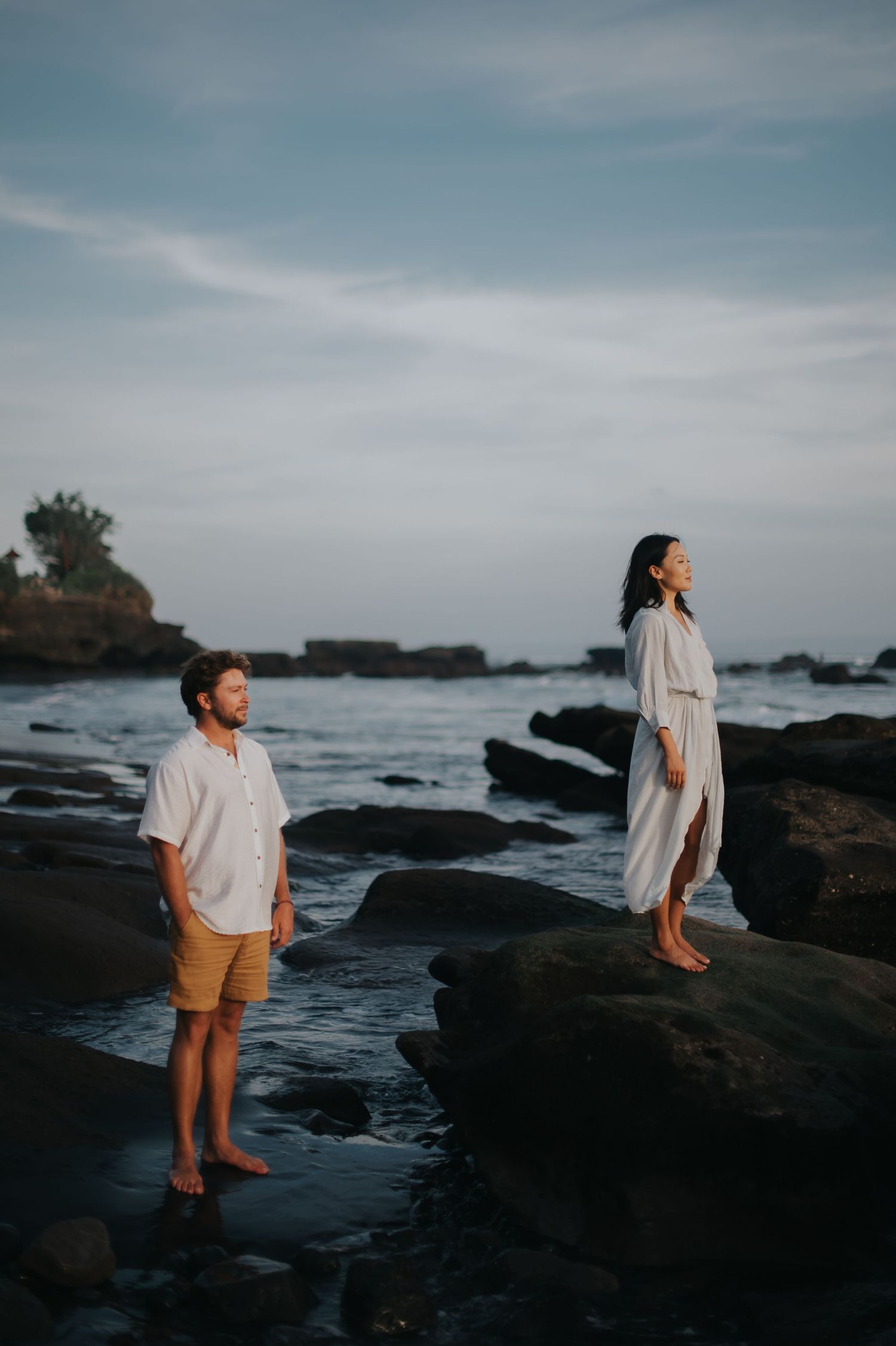 Two people stand on rocky coastal cliffs at sunset, wearing light colored clothing as waves crash against the shore.