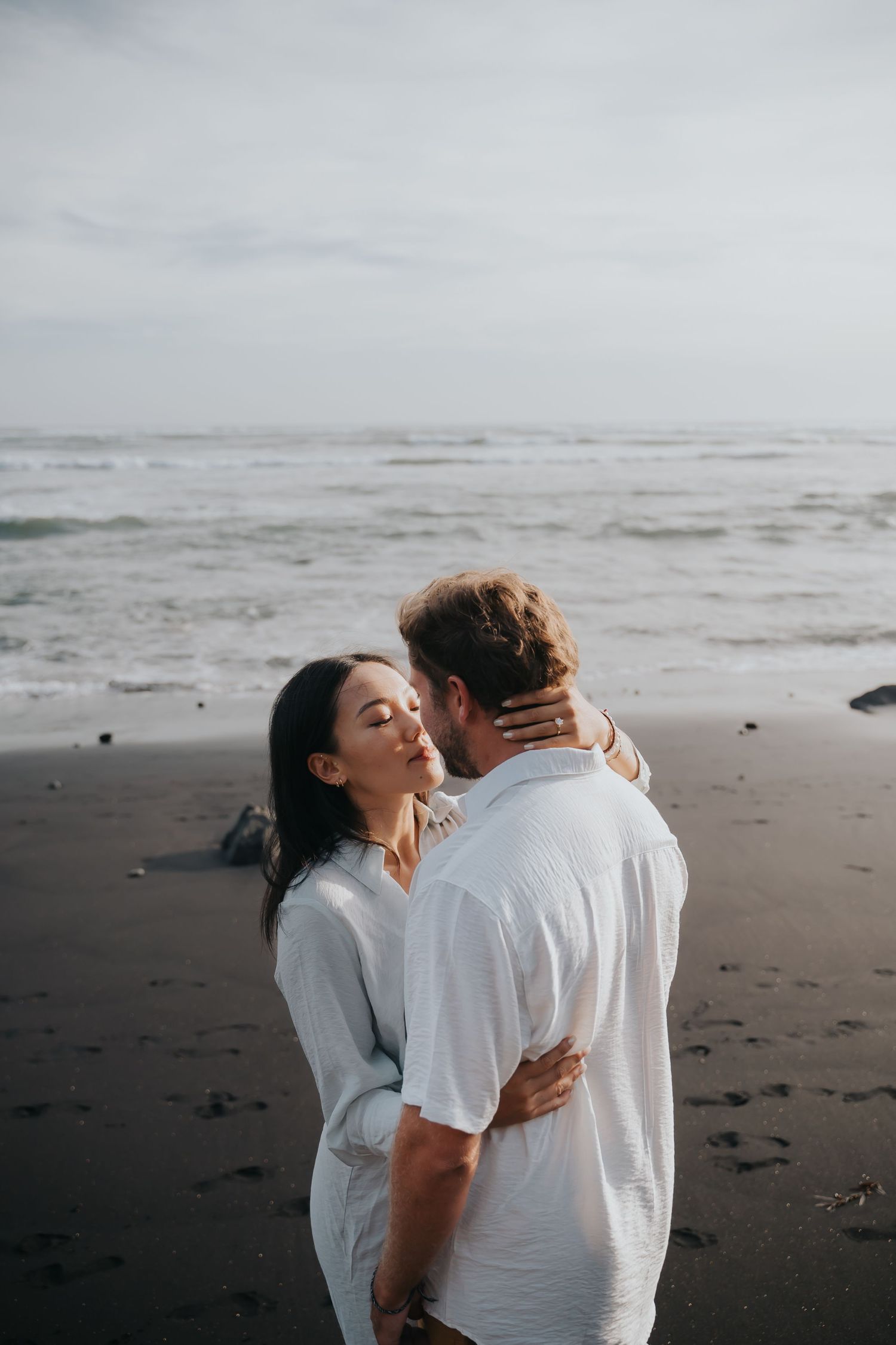 A couple embraces lovingly on a moody beach with dark sand and overcast skies.