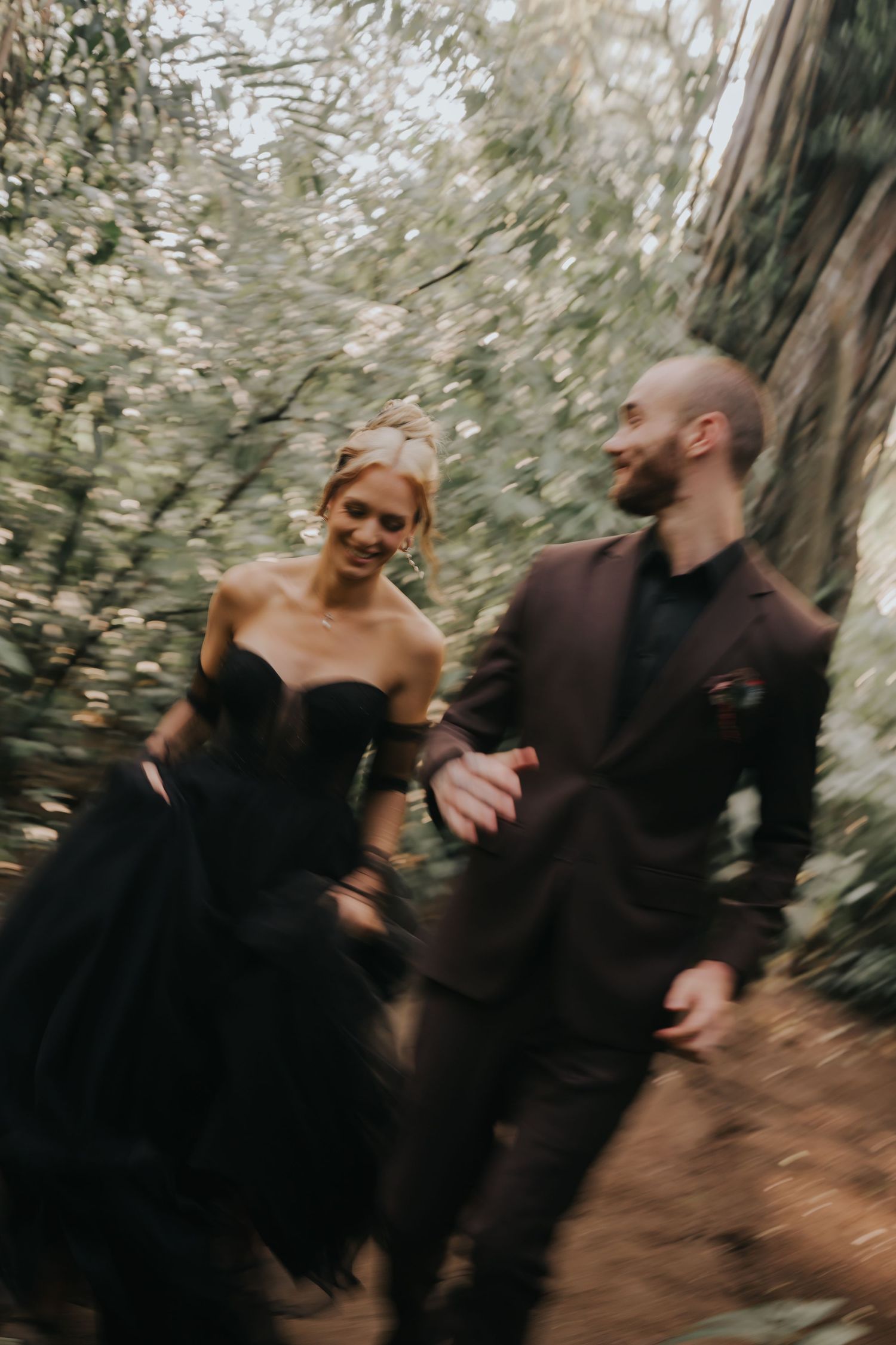 A couple in black attire sharing a playful moment in a forest setting.