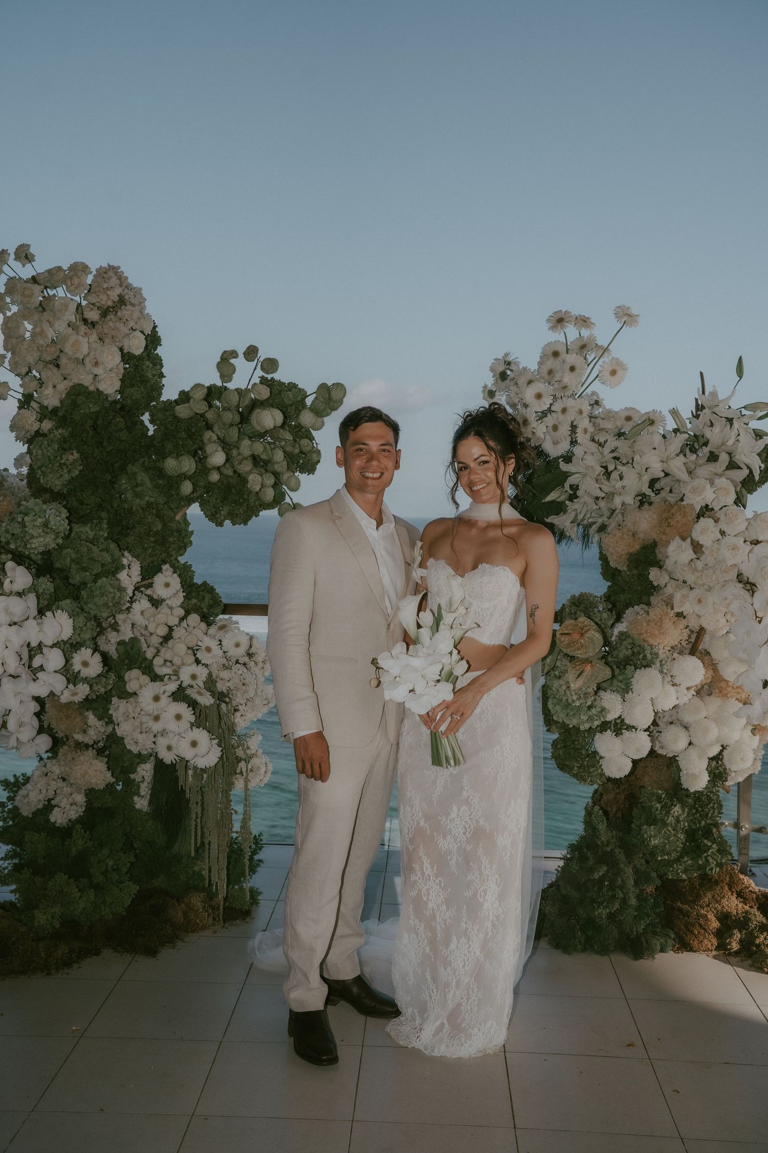 A couple celebrates their wedding day in front of white floral arrangements with the ocean in the background.