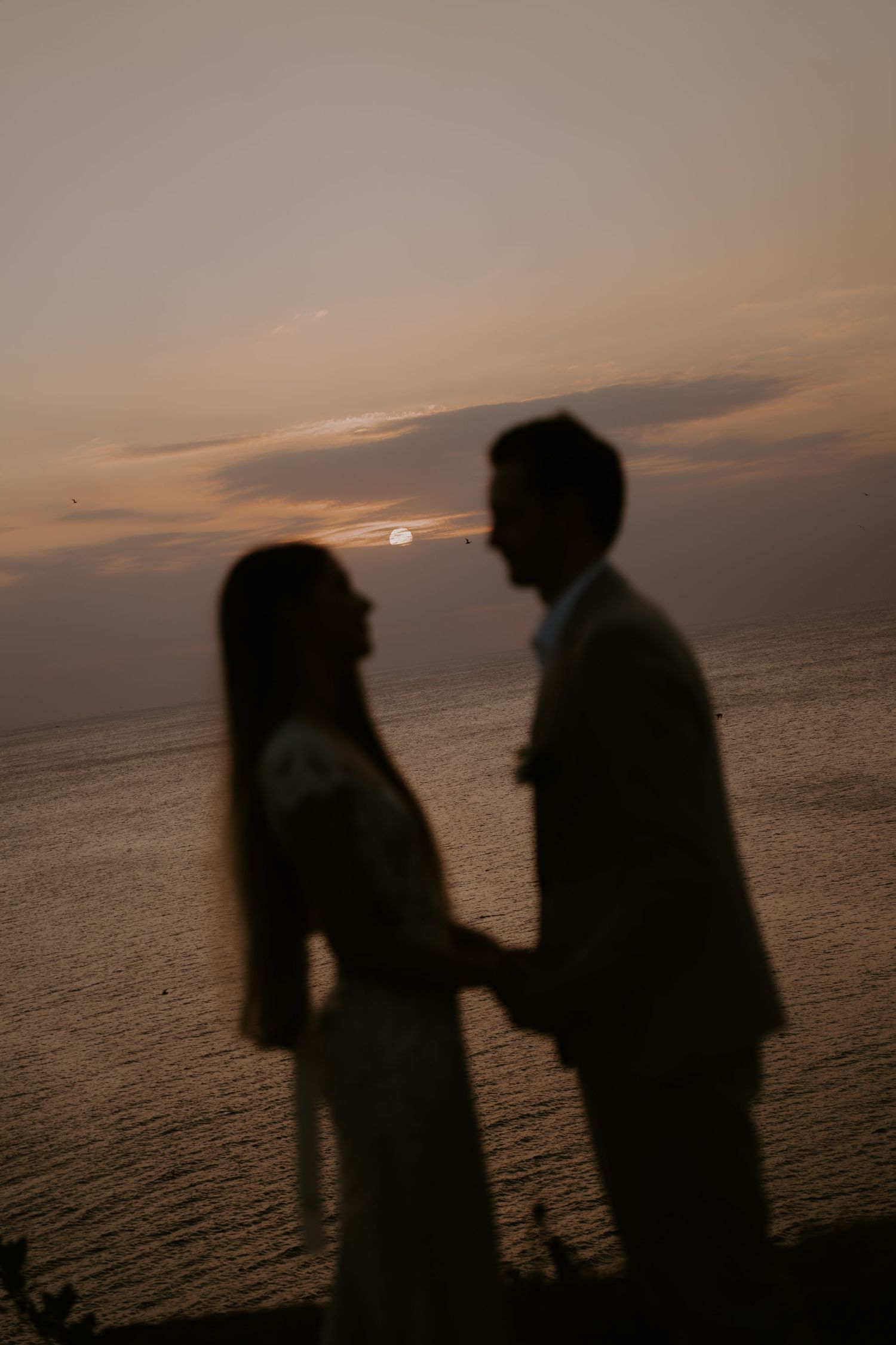 Silhouettes of two people stand together against a sunset over the ocean creating a romantic atmosphere.